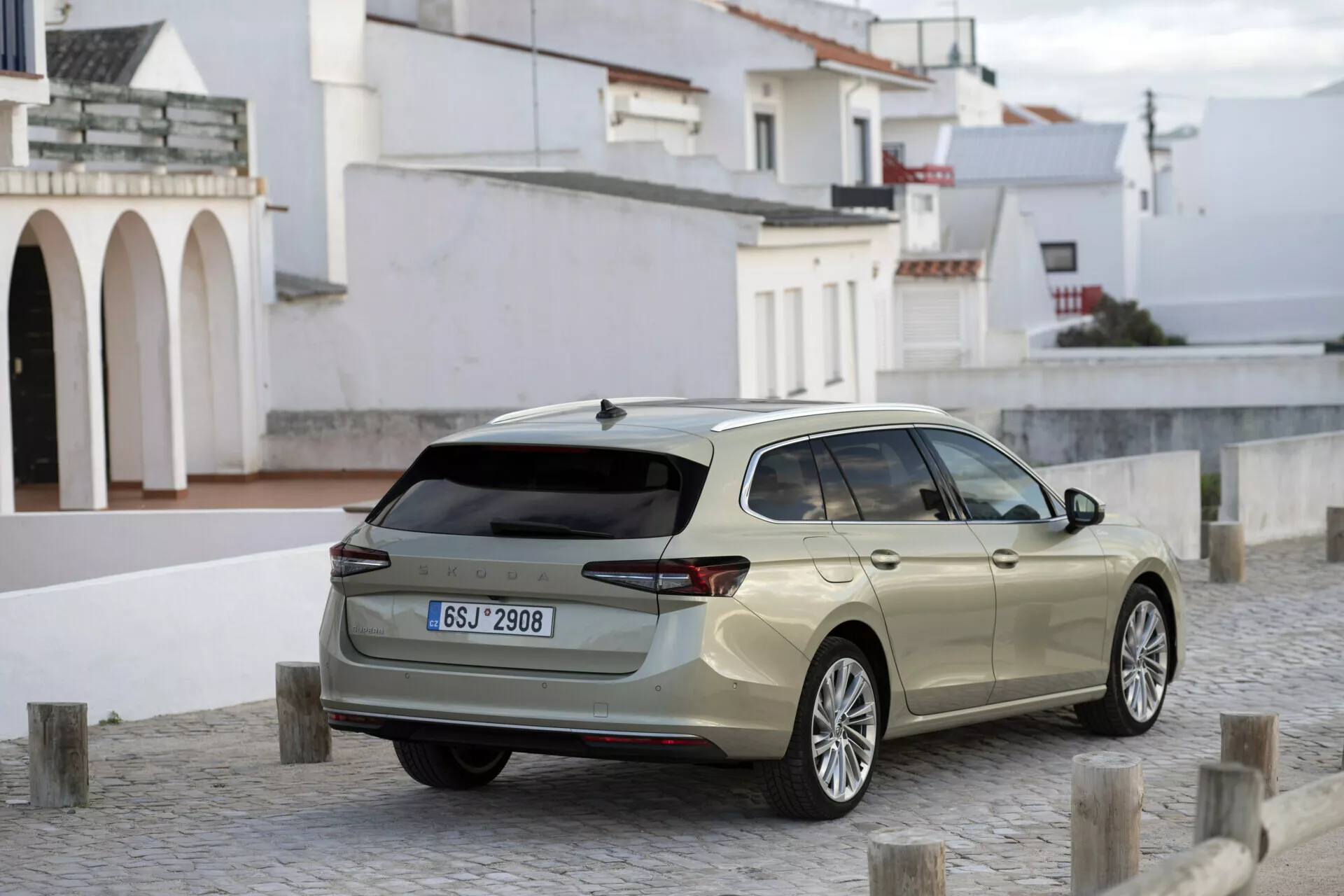 Bildens visar en modern Skoda Superb 2024 kombi från en trekvarts bakre perspektiv, parkerad på en kullerstensyta med flera korta stolpar som fungerar som barriärer. Kännetecken inkluderar moderna LED-bakljus, stor bakre vindruta med torkarblad, parkeringssensorer i bakre stötfångaren, särskiljande aluminiumfälgar, silverfärgade takräcken och en 'hajfena' antenn. Sidprofilen avslöjar en förlängd bakre del, vilket indikerar gott om lastutrymme. Naturligt dagsljus framhäver fordonets design och användbarhet i en möjlig småstads- eller bymiljö.