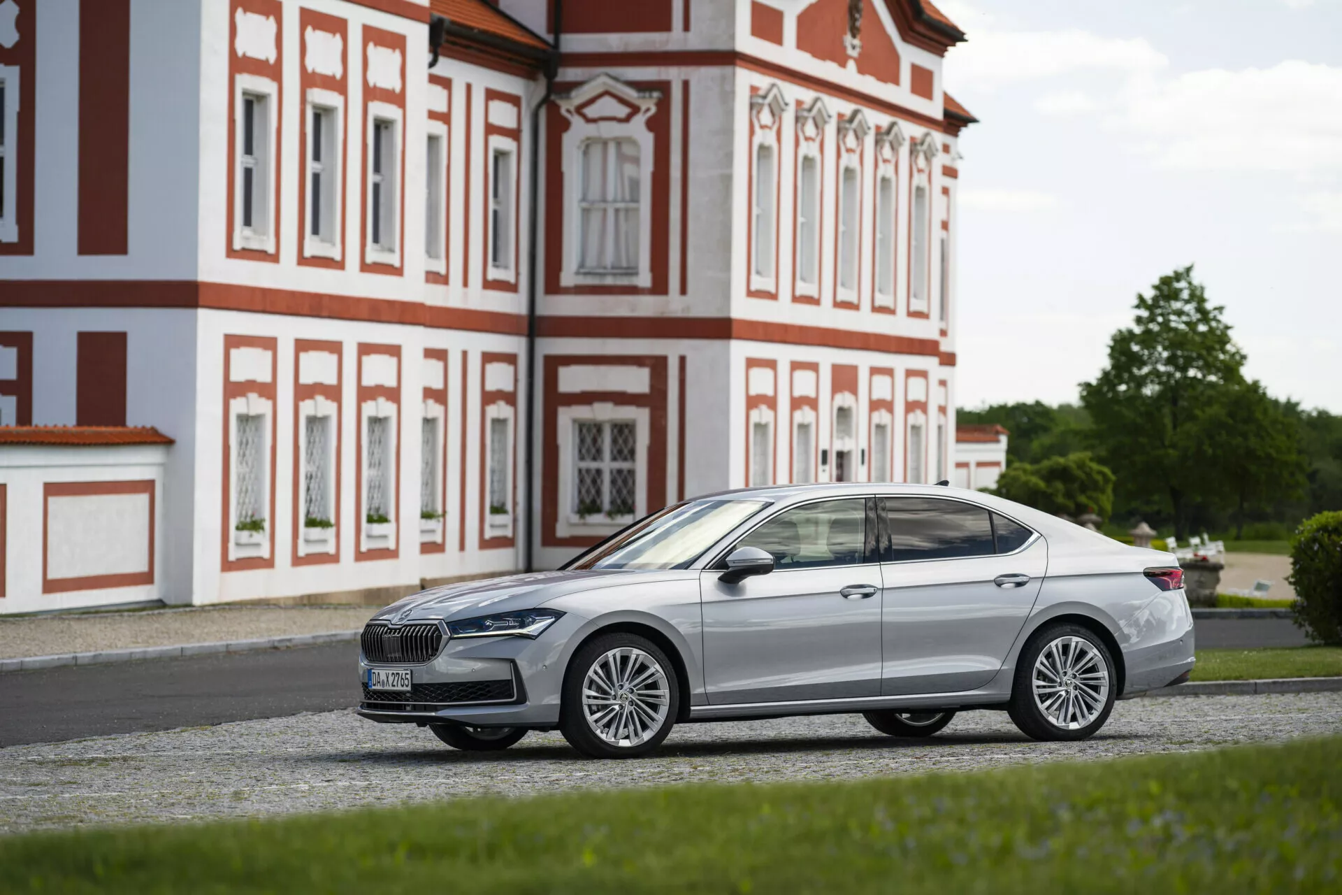 Sidoprofil av en modern sedan parkerad på grus, fokuserande på passagerarsidan, tagen från en låg vinkel. Bilen, placerad parallellt med kameran, visar sin eleganta design, kromade detaljer, stiliga lättmetallfälgar och en betydande frontgrill med en framträdande logotyp. Den släta, aerodynamiska silhuetten med definierade linjer, nedfällda sidospeglar och infällda dörrhandtag tyder på en avancerad nyckelfri åtkomst. Tonade rutor antyder integritetsskydd eller UV-skydd. En barockbyggnad i bakgrunden kontrasterar mot bilens modernitet.
