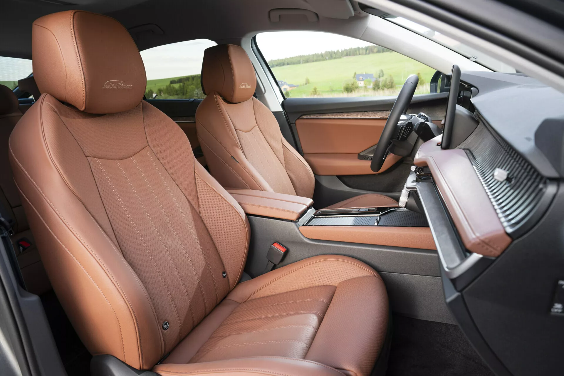 Bilden visar interiören av en 2024 års Skoda Superb Limousine, med fokus på området för föraren och passageraren framifrån, sett från passagerarsidan. Den visar lyxiga bruna lädersäten med ett lyxemblem i relief, en multifunktionell läderklädd ratt med kontrollknappar och design med platt botten, en instrumentpanel med kolfiberliknande trim med möjlighet till ambient belysning och en mittkonsol som innehåller en växellåda, mugghållare och förvaringsfack. Dörrens interiör matchar de bruna lädersätena med borstade metalldetaljer. Golvet är heltäckningsmatta för extra komfort och ljudisolering, vilket betonar bilens lyxiga och tekniskt avancerade egenskaper.