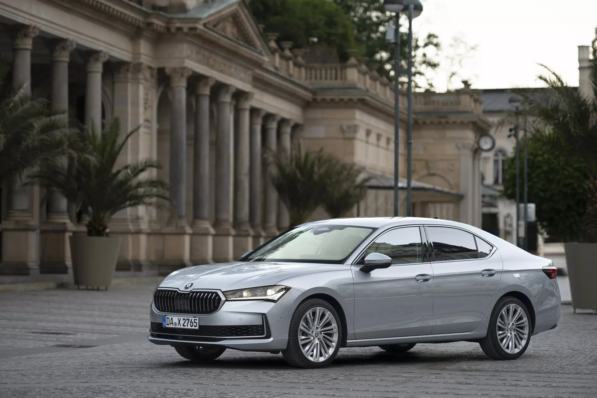 Den här bilden visar den moderna Skoda Superb 2024-sedan parkerad på ett asfalterat öppet område, troligtvis ett torg eller en innergård, med en klassiskt utformad byggnad med kolonner i bakgrunden, vilket skapar en formell miljö som kan associeras med regeringsbyggnader eller kulturplatser. Bilen är fotograferad från en trekvarts vinkel framifrån, något vinklad mot förarsidan, vilket ger en klar vy av både fram- och sidoprofiler. Designinslag som framhävs inkluderar den framträdande grillen med ett specifikt mönster, de eleganta horisontella LED-strålkastarna, flerspakiga lättmetallfälgar, aerodynamiska karosslinjer, sidospegeln med inbyggda blinkers och den metalliska lacken. Märkesemblemet på grillen och eventuellt modellbeteckning bakåt understryker fordonets premiumposition.