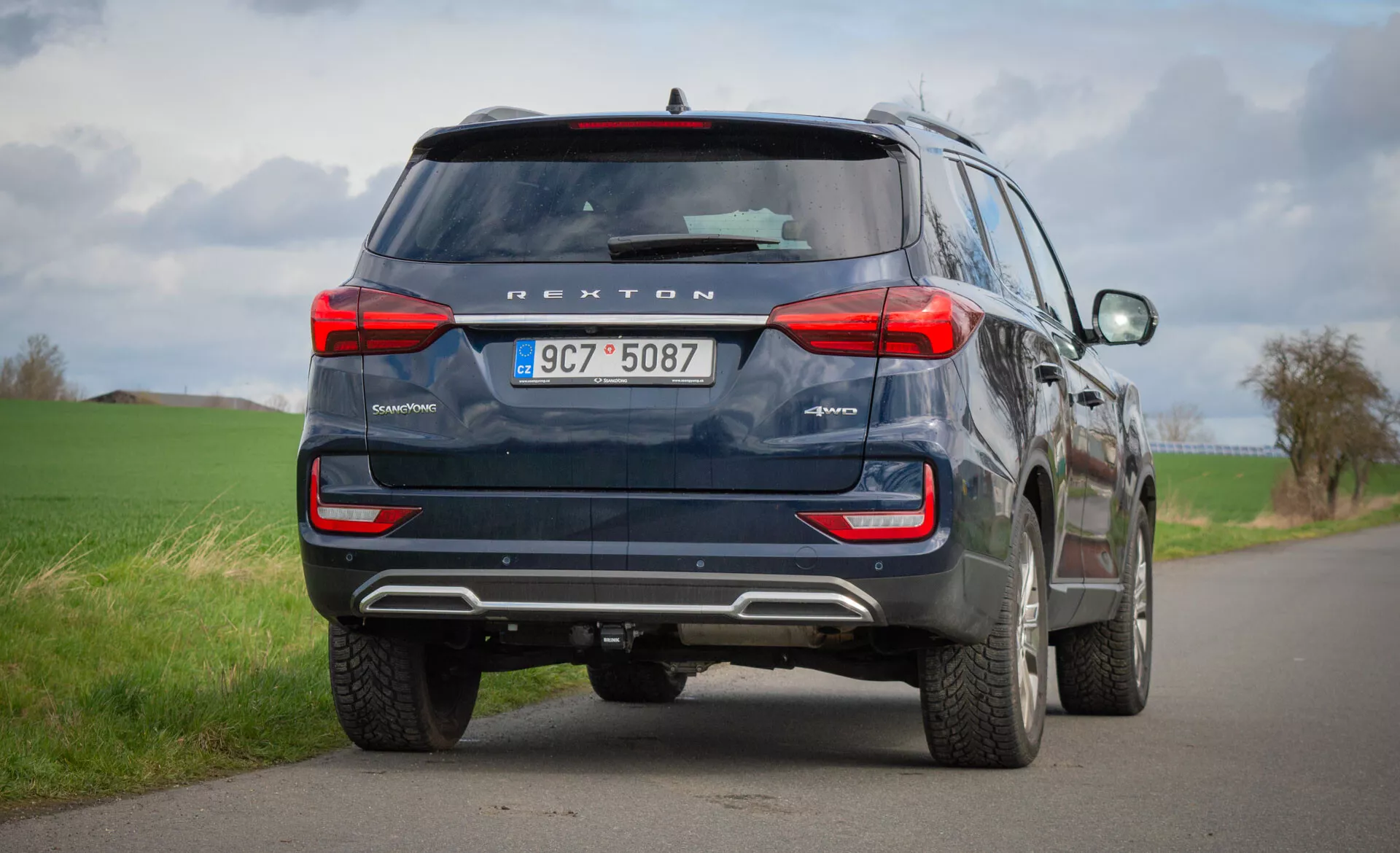 Baksida av en blå SsangYong Rexton SUV 2021 parkerad på en landsväg med två körfält, som framhäver dess designfunktioner som bakljus, bakruta, avgasrör och säkerhetsteknologi inklusive backkamera och parkeringssensorer, under en molnig himmel.