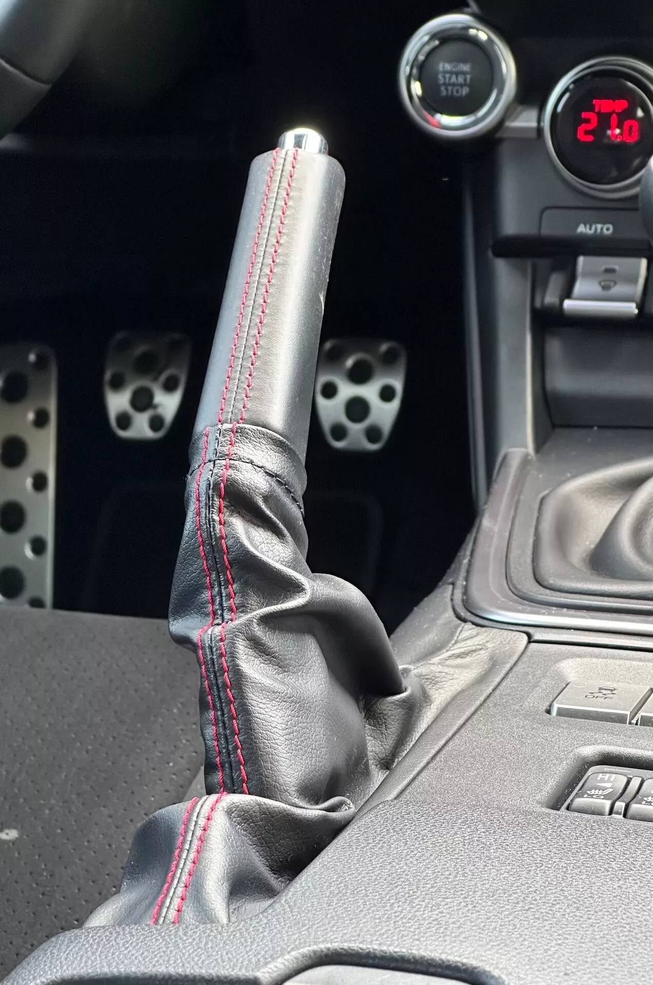 Närbild av mittkonsolen i Subaru BRZ 2023 som framhäver den metalliska växelspaksknoppen med röda kontrastsömmar på skinnstöveln, en digital mätare som visar '21', sportiga aluminiumspedaler med borrade hål och tekniska funktioner såsom knappar för elektronisk stabilitetskontroll.