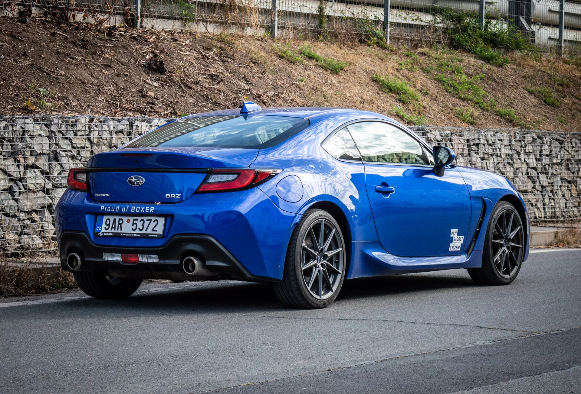 2023 Subaru BRZ sportcoupé i metalliskt blått parkerad på en lutning, visar sin bakre tre fjärdedels vy som betonar fordonets sidoprofil, bakre kurvatur, sportiga legeringshjul, dubbla avgasutblås, läppspoiler och 'Proud of Boxer' motorbadge.