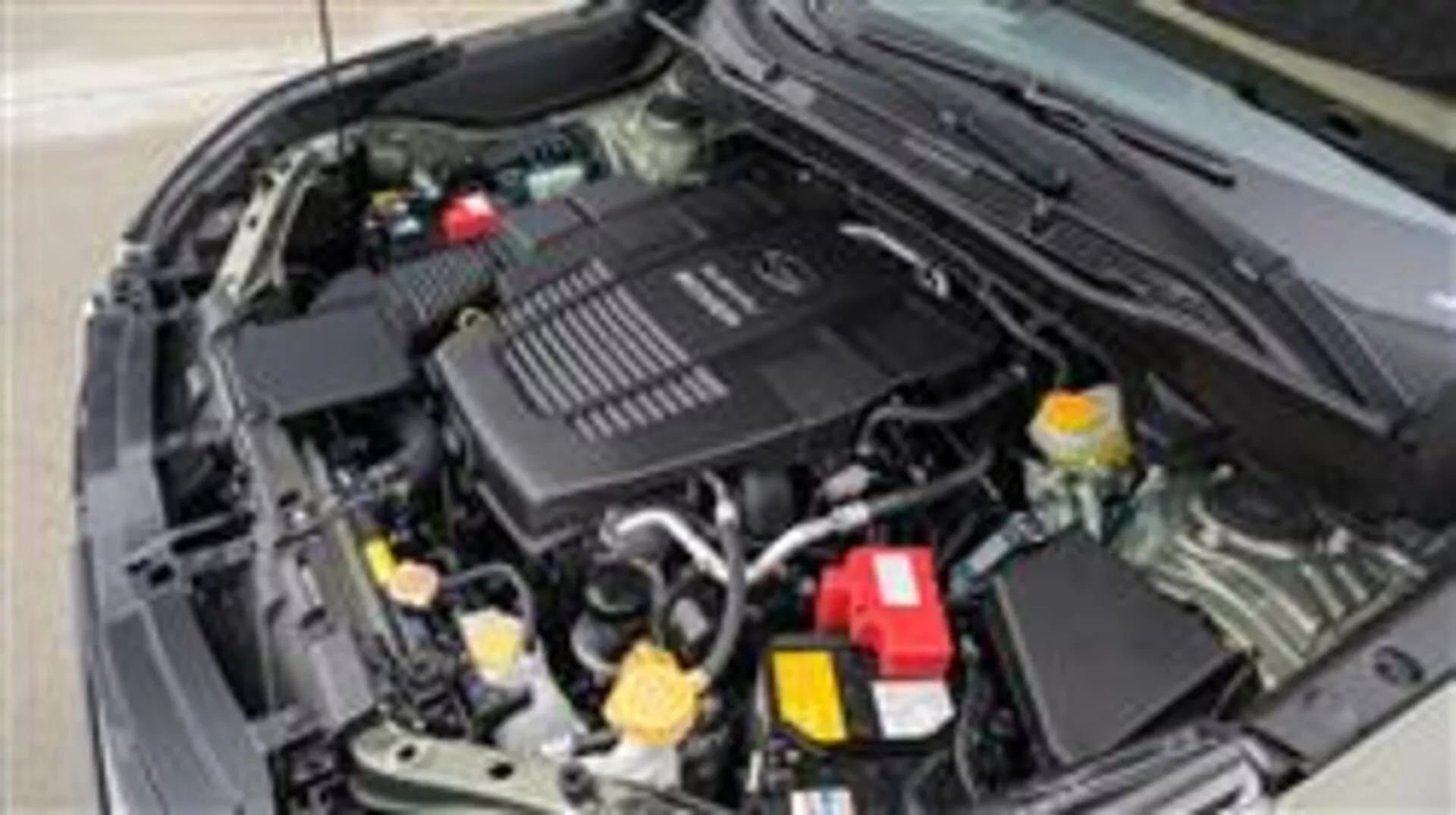 Närbild av motorutrymmet i Subaru Forester 2015 som visar batteri, vätskebehållare, luftfilterlåda, olika rör och slangar, mekaniska delar och motorlock