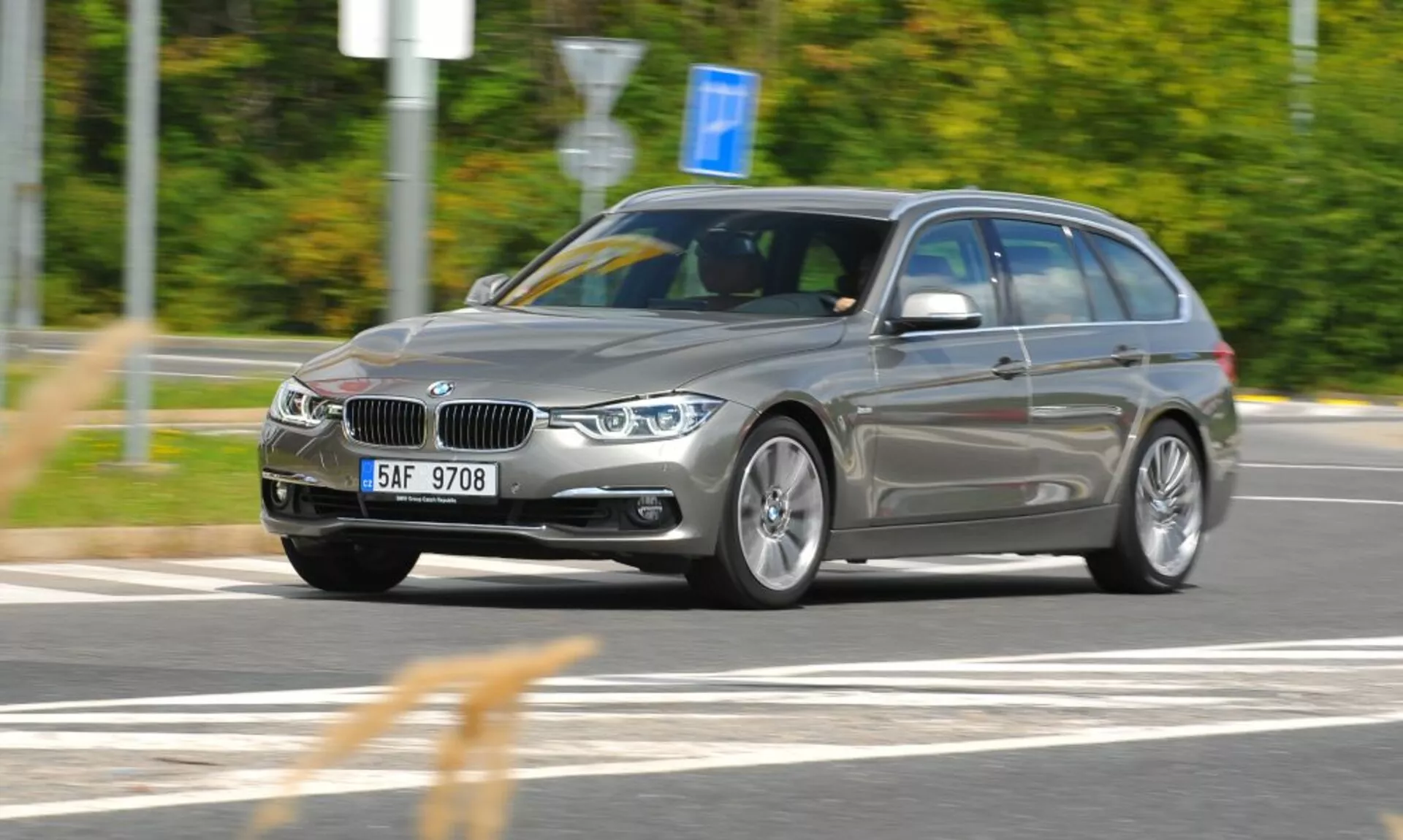 Dynamisk tre fjärdedels framifrån-vy av en silver BMW kombi i rörelse med distinkt njurformad grill och emblem, fångad från en lätt höjd vinkel indikerande hastighet och rörelse, fokuserad på design och tekniska egenskaper såsom vinklade strålkastare, legeringshjul och takräcken i en rörelseoskärpa utomhusmiljö.