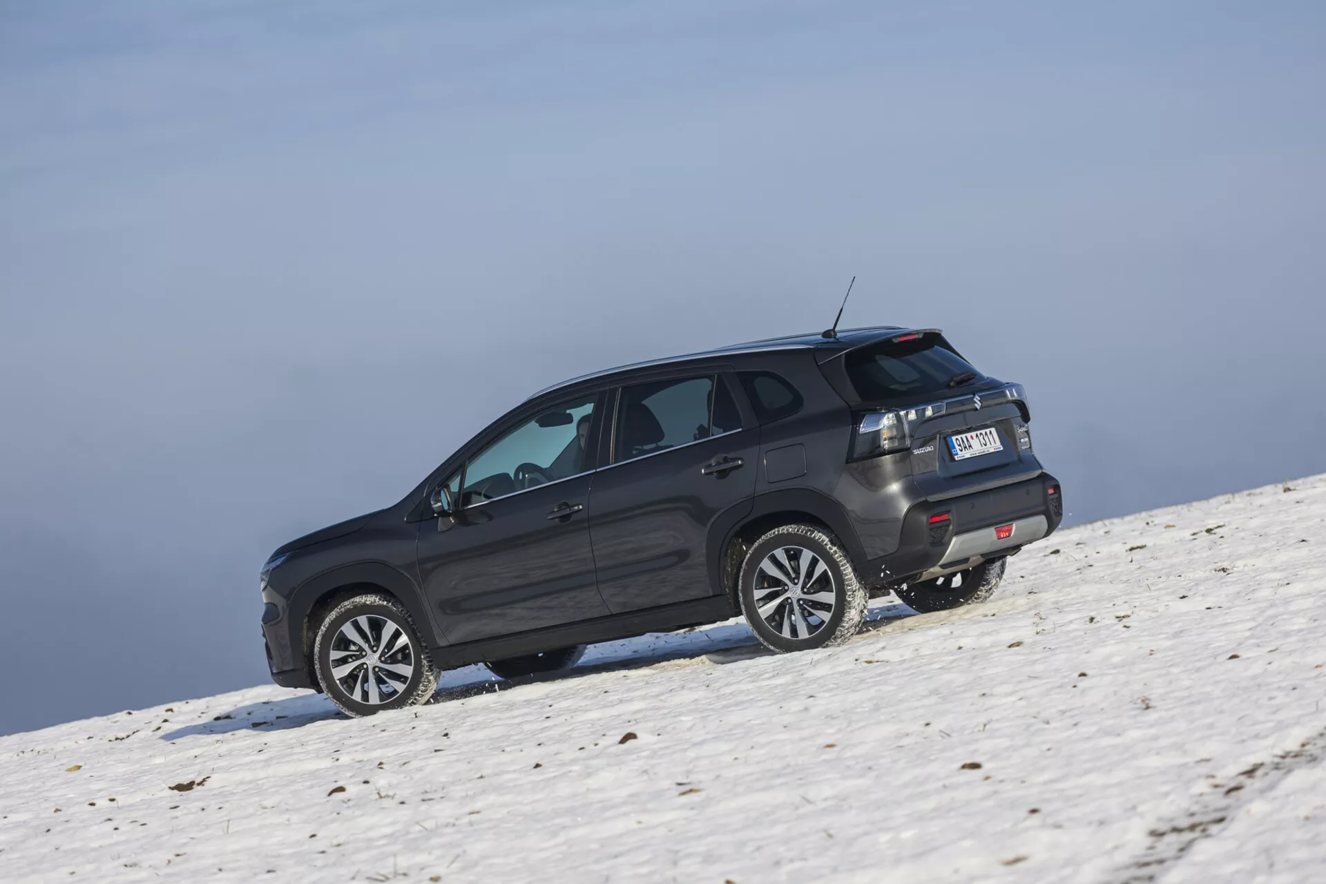 SUV Suzuki S-Cross 2022 i mörk nyans placerad på en snötäckt sluttning fångad från en sned bakre vinkel som framhäver dess sida och bakprofil, stora lättmetallhjul, tonade rutor med avfrostare, LED-bakljus, takspoiler, och potentiella offroad-förmågor under klara himmelsförhållanden.