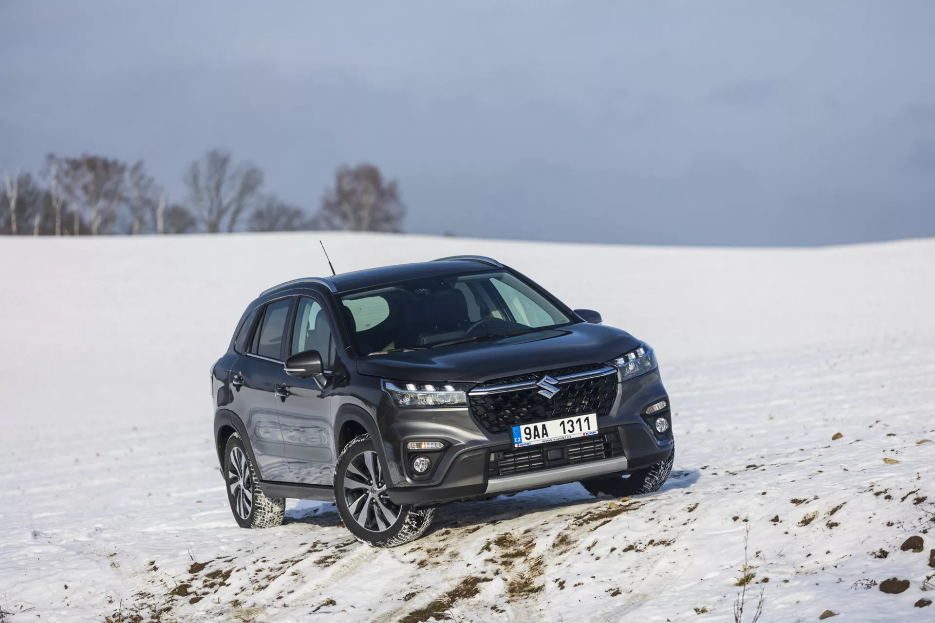 Mörkfärgad kompakt SUV Suzuki S-Cross årsmodell 2022 parkerad i ett snötäckt landskap, visar en främre och sidoprofil vid 45 graders vinkel, med en snygg grill med metallisk finish, förlängda LED-strålkastare, flerspakiga aluminiumfälgar och polerad kromad fönsterlist som indikerar en lyxig utrustningsnivå och potentiell terrängförmåga