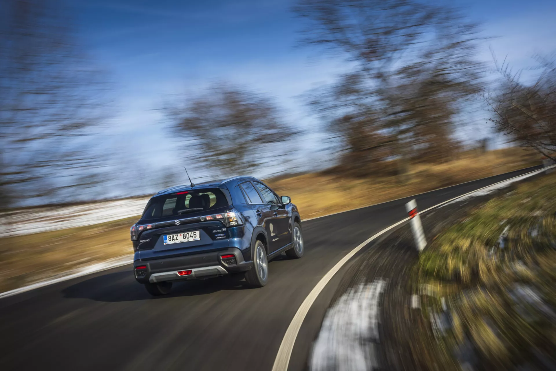 Modernt SUV Suzuki S-Cross 2022 i rörelse, visar bakre tre kvarts profilvy med snurrande hjul på en klar dag