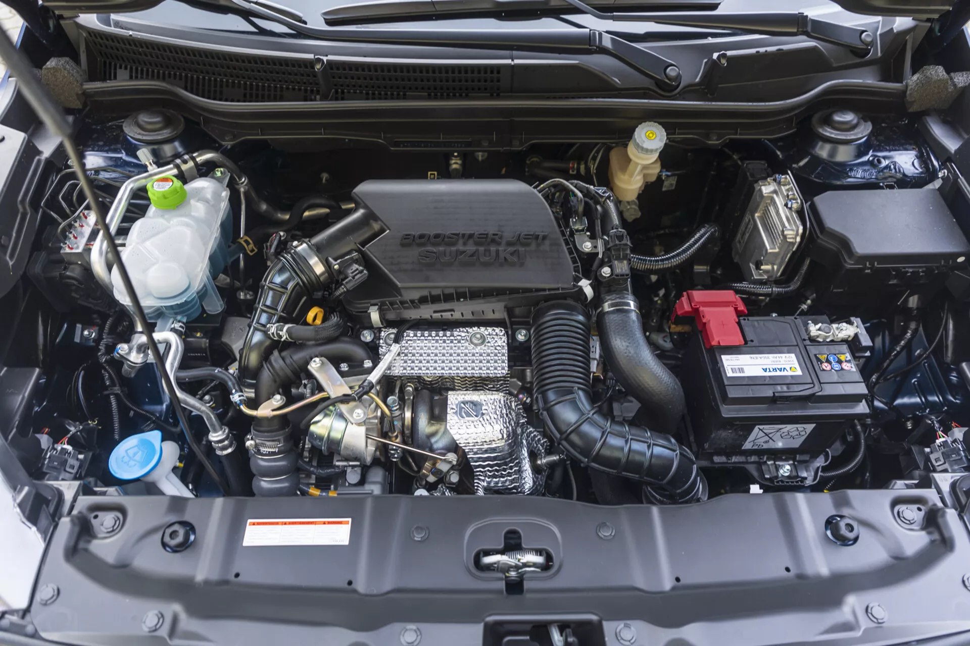 2022 Suzuki S-Cross motorutrymme som visar en front-vy med synliga komponenter som motorblock, cylindertopp, insugningsmunstycke och tekniska funktioner inklusive turbo och variabel ventiltiming, tillsammans med Suzuki-branding på motorkåpan, i nyskick.