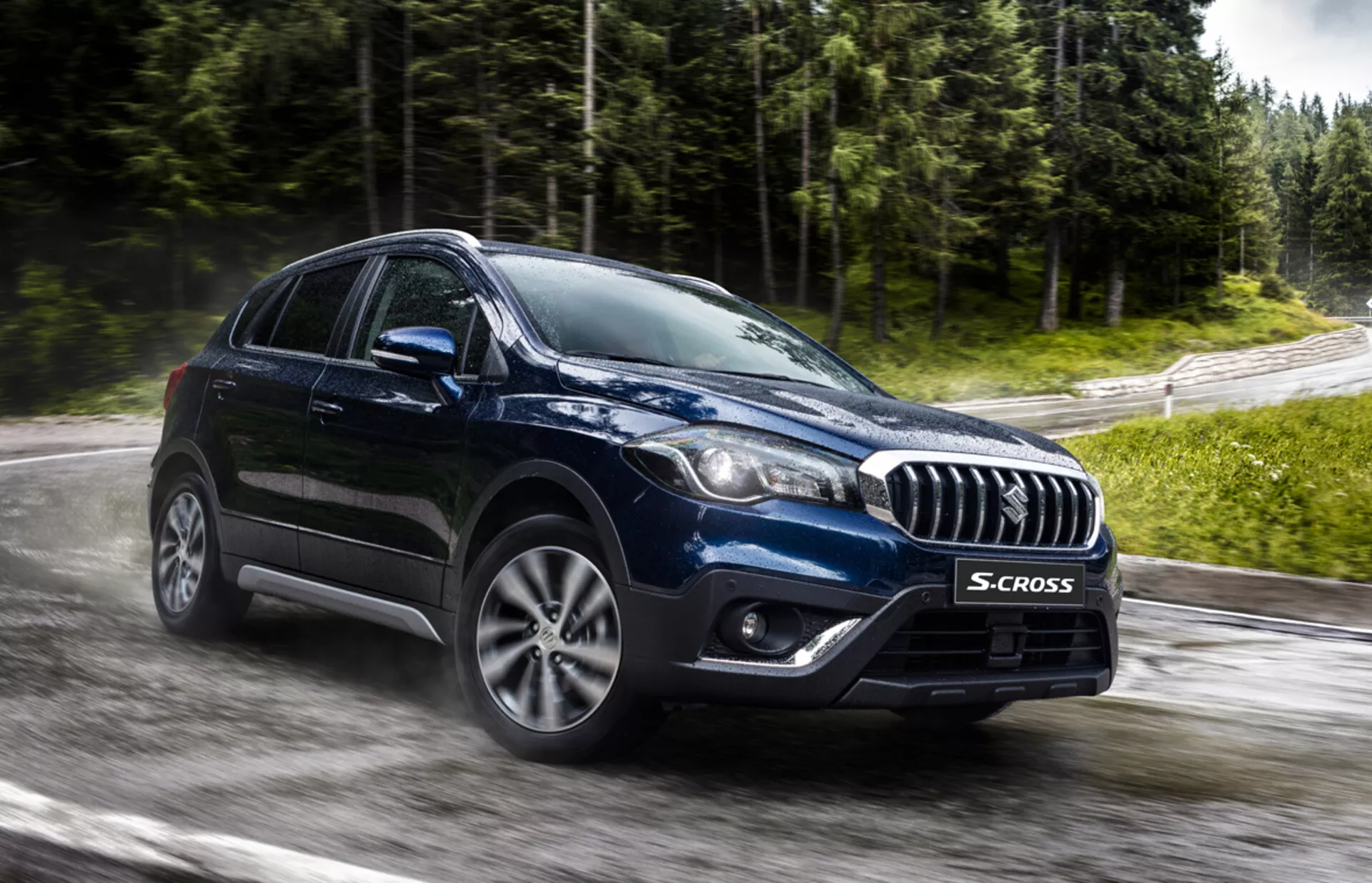 Suzuki S-Cross 2022 fångad från ett frontal tre kvarts vinkel som framhäver dess kromaccentuerade grill, moderna LED- eller HID-strålkastare, flerspakiga legeringshjul och robust crossover-karossform på en våt väg omgiven av grönska, vilket antyder avancerad teknologi och allväderskapacitet.