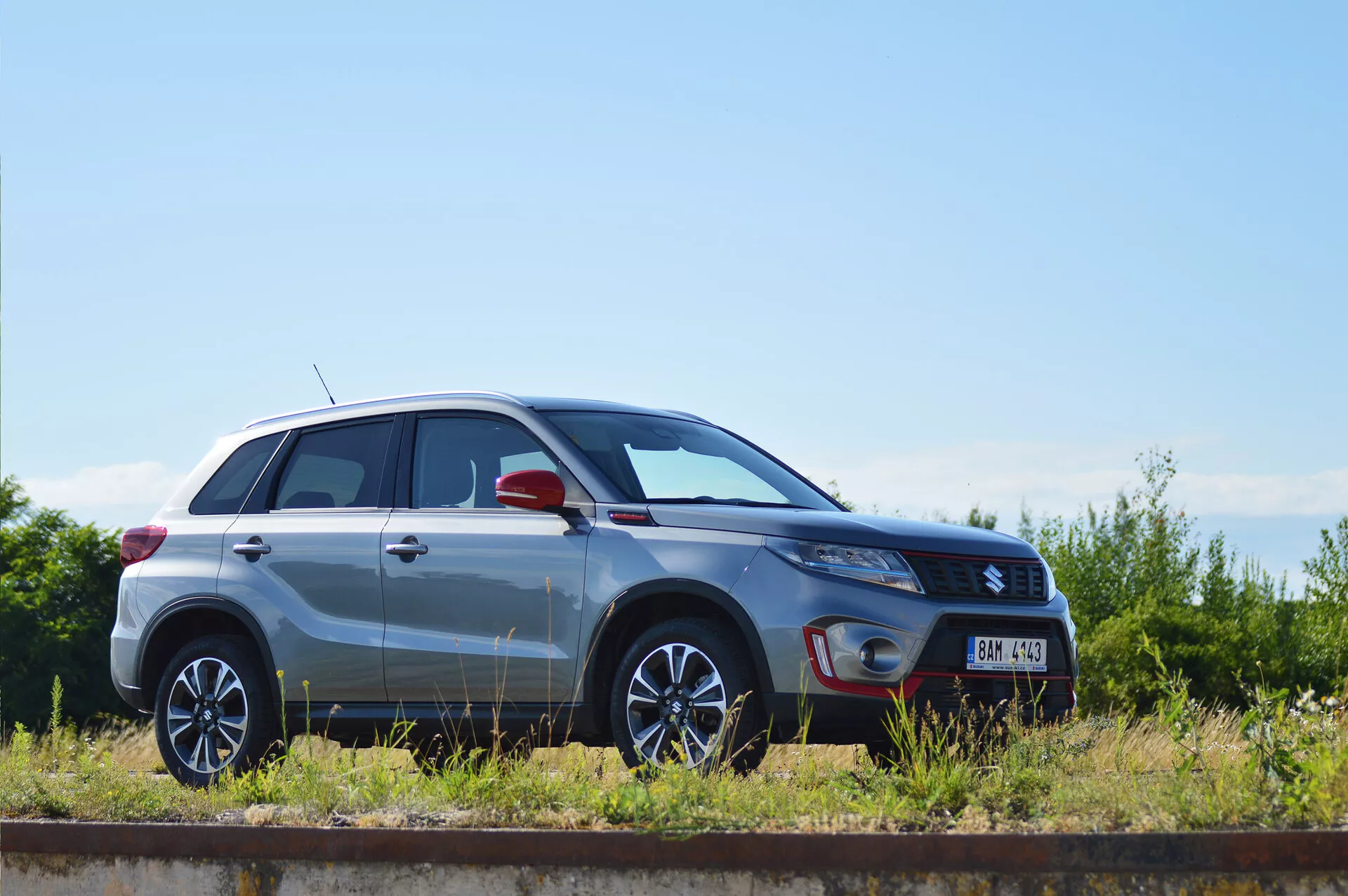 Sidoprofilvy av en silverfärgad Suzuki Vitara compact SUV från 2018, parkerad parallellt med ett järnvägsspår på en solig dag. Designlinjerna, de flerfärgade fälgarna, metallisk lackfinish, stängda dörrar och fönster, strålkastarnas form, sidospeglar med potentiellt integrerade blinkers och trimbadge är tydligt synliga, vilket visar fordonets design och teknologiska egenskaper.
