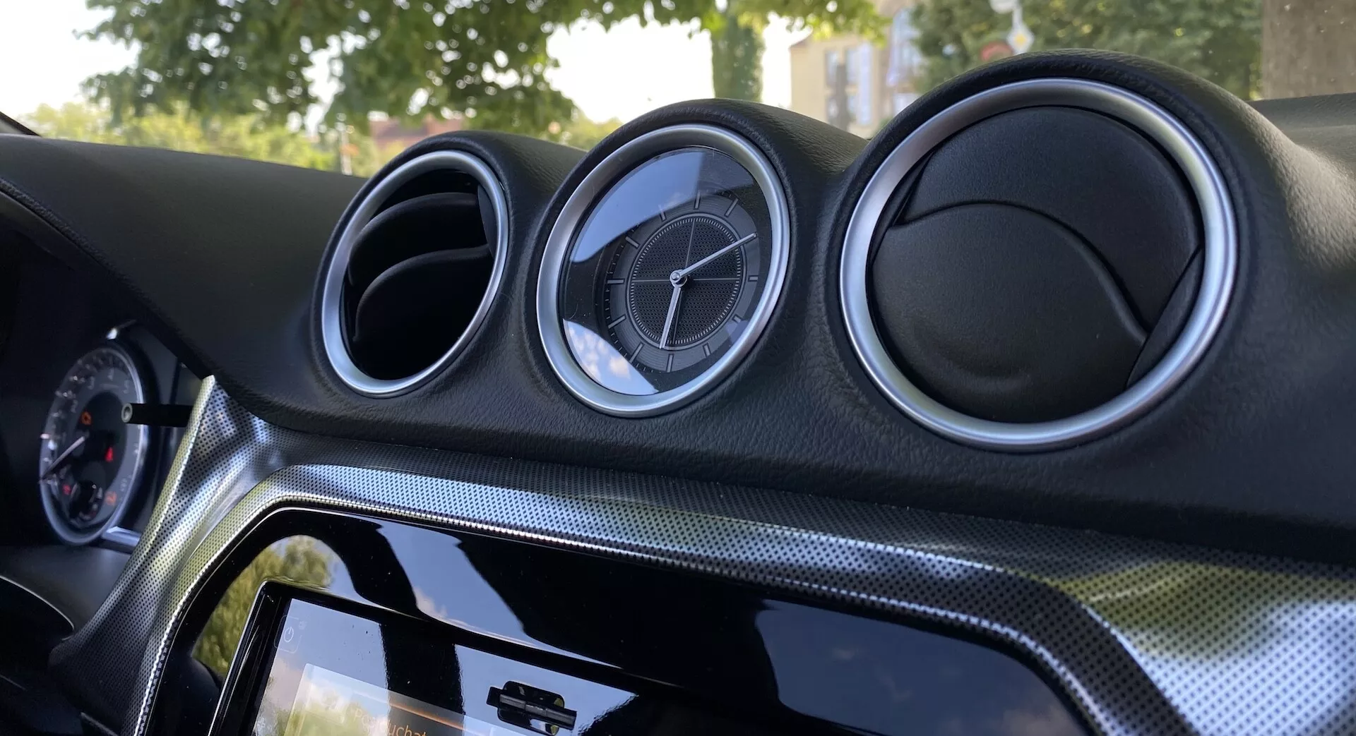 Närbild på interiören i Suzuki Vitara 2018 med fokus på den centrala instrumentpanelen. Instrumentpanelen har tre cirkulära luftventiler med metallisk utseende och tillverkarens emblem, en pekskärmsinfotainmentsystem med blank svart kant, material med kolfiberutseende och omgivande belysning för förbättrad estetik.