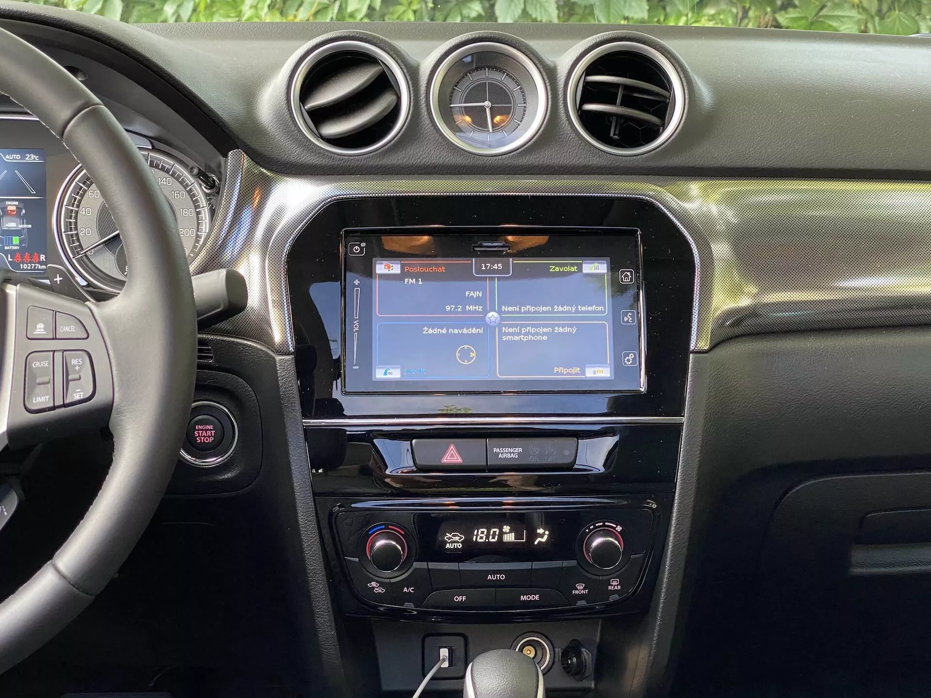 Interiörvy av Suzuki Vitara 2018 med fokus på den centrala instrumentbrädan. Sektionen visar en delvis synlig ratt till vänster med flera kontrollknappar, troligtvis för ljud, handsfree-samtal och farthållare. Den centrala konsolen visar en infotainmentskärm, som troligen visar navigering eller en menyer, flankerad av vertikala luftutsläpp och toppad med tre cirkulära luftventiler. Under pekskärmen finns en rad knappar och rattar för snabb åtkomst till infotainmentsystemet och fordonsinställningar. Klimatkontrollerna är placerade längre ner och antyder ett klimatsystem med två zoner. Instrumentbrädan har ett mjukt material med synliga sömmar och en rund analog klocka är monterad mellan de centrala cirkulära luftutsläppen. Synligheten är fokuserad på instrumentbrädans funktioner, utan fönster eller vindrutor i sikte.