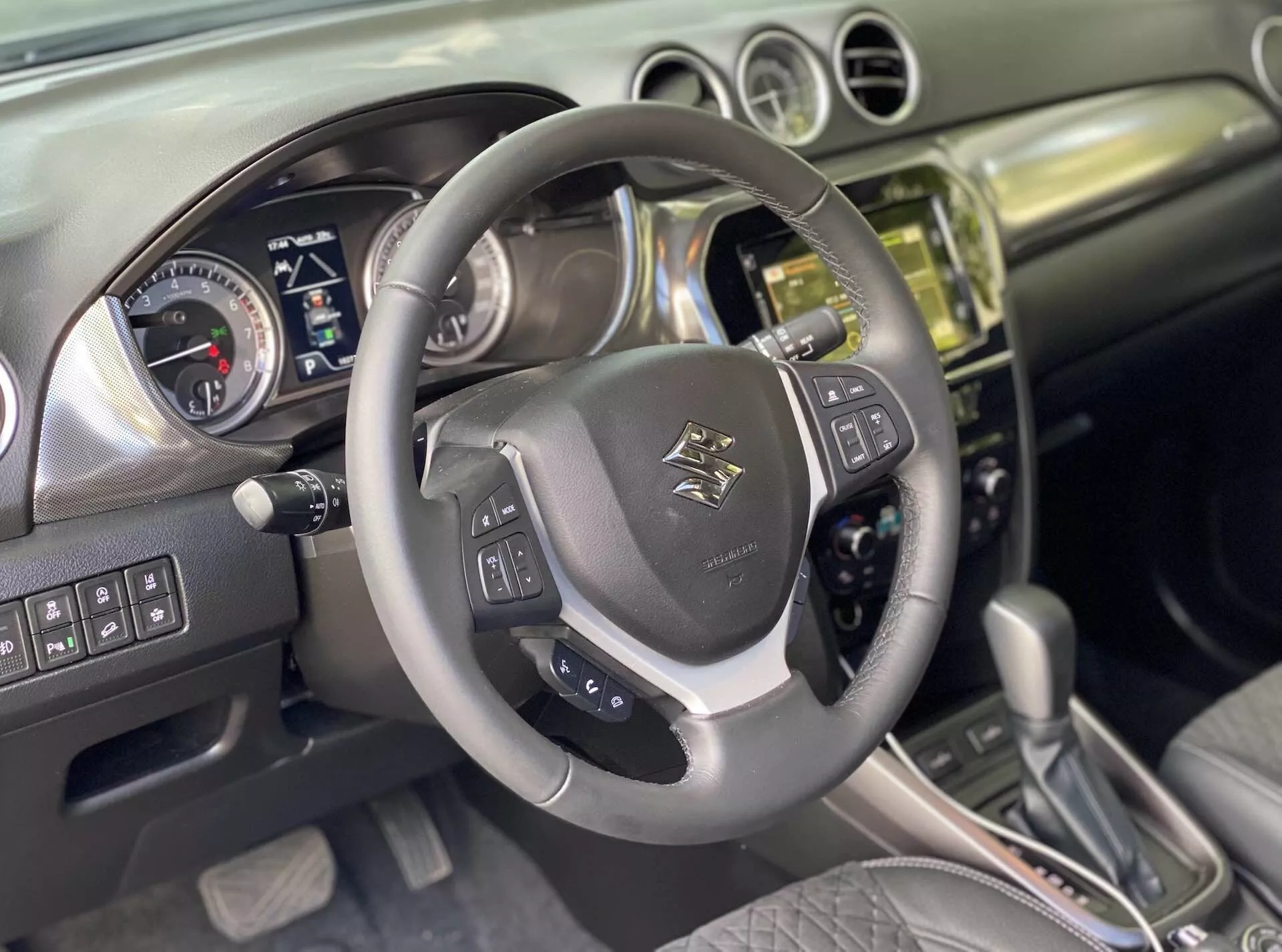Interiör i en Suzuki Vitara 2018 sett från förarens perspektiv. Närbild på en multifunktionell ratt klädd i läder med emblem i mitten, med monterade reglage för ljud, röststyrning och farthållare med metallaccenter. Bakom ratten finns en instrumentbräda med analoga mätare för hastighetsmätare och varvräknare, och en digital skärm för fordonsinformation som indikerar att bilen är avstängd eller i tillbehörsläge. Mittkonsolen visar en stapel i metall eller krom med ett pekskärms-infotainmentsystem, knappar och reglage för klimat- och fordonskontroller. En automatisk växelspak med läderdetaljer antyder olika körlägen. Interiören kombinerar läder och borstad metall, vilket speglar en modern design. Avancerade funktioner som förmodligen inkluderar Bluetooth, navigation, parkeringsassistenter och förarassistanssystem.