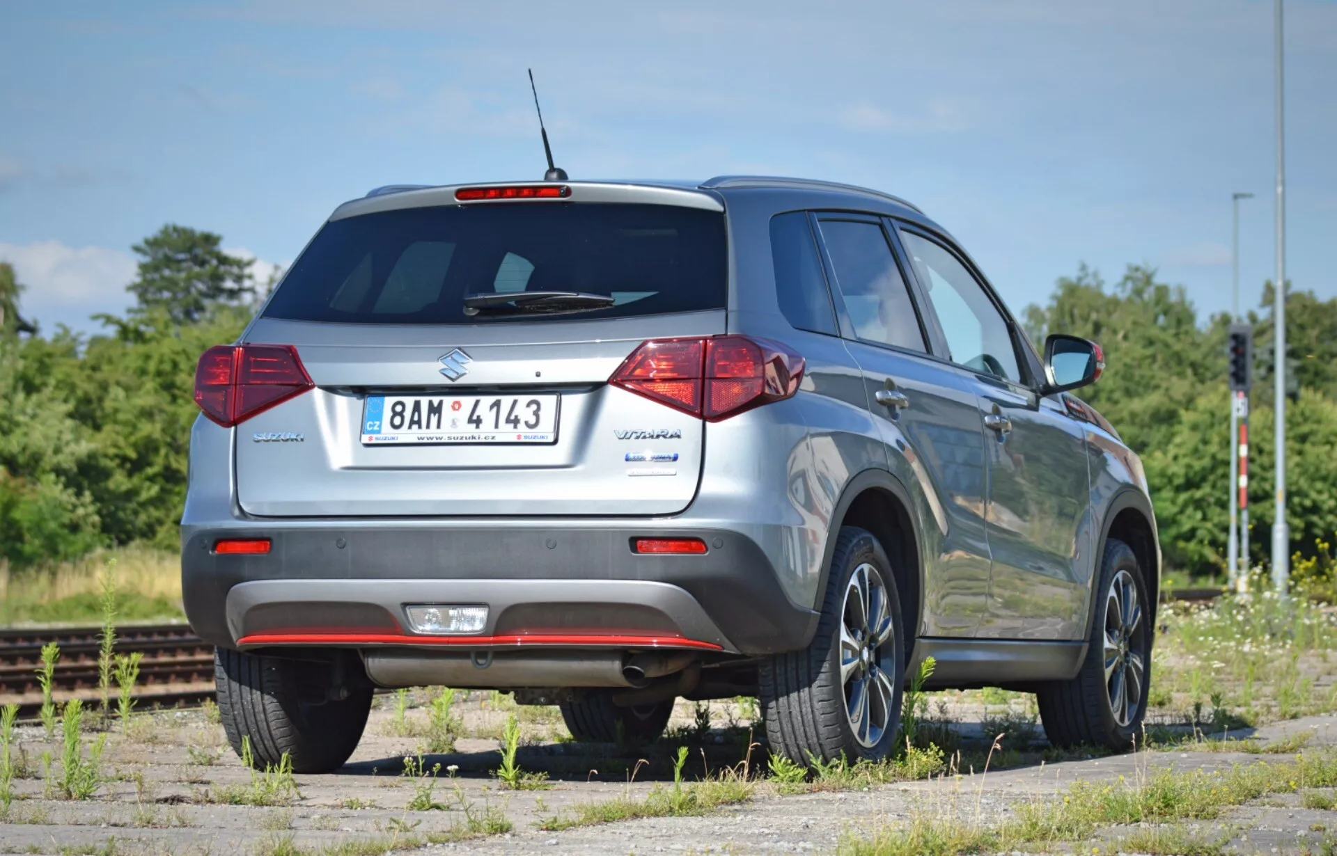 Bakgrund av en Suzuki Vitara 2018 SUV från en låg vinkel, som betonar dess höjd och markfrigång. Positionerad i en 45-graders vinkel, visar bilden bakluckan med modellmärke, bakruta med vindrutetorkare, traditionella halogenbakljus, reflekterande bakre stötfångare och synligt avgasrör. Högersidans profil innehåller passagerardörren bak, sidorutor, takräcken och standard aluminiumfälgar. Inga synliga blinkers på sidospeglarna. Fordonet är parkerat på en dammig yta nära järnvägsspår, i ett torrt, soligt område med grönska och blå himmel.