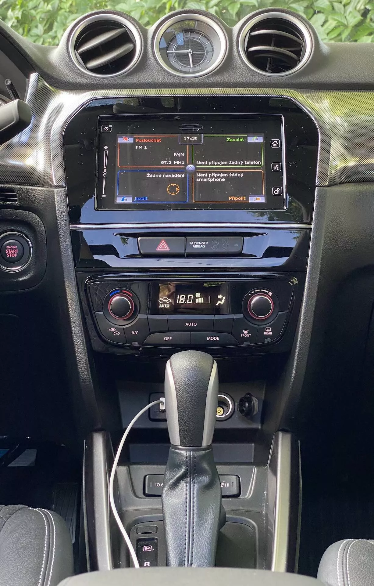 Djupgående vy över interiören i en Suzuki Vitara 2018, med fokus på mittkonsolen och instrumentbrädan från framsätets perspektiv. Bilden framhäver bilens infotainmentskärm som visar en meny, ett klimatsystem med två zoner och digitala avläsningar samt en växelspak för automatisk växellåda. Interiören har en mörkfärgad finish med en blandning av matta och glansiga ytor, vilket illustrerar materialkvalitet och modern bilteknik.