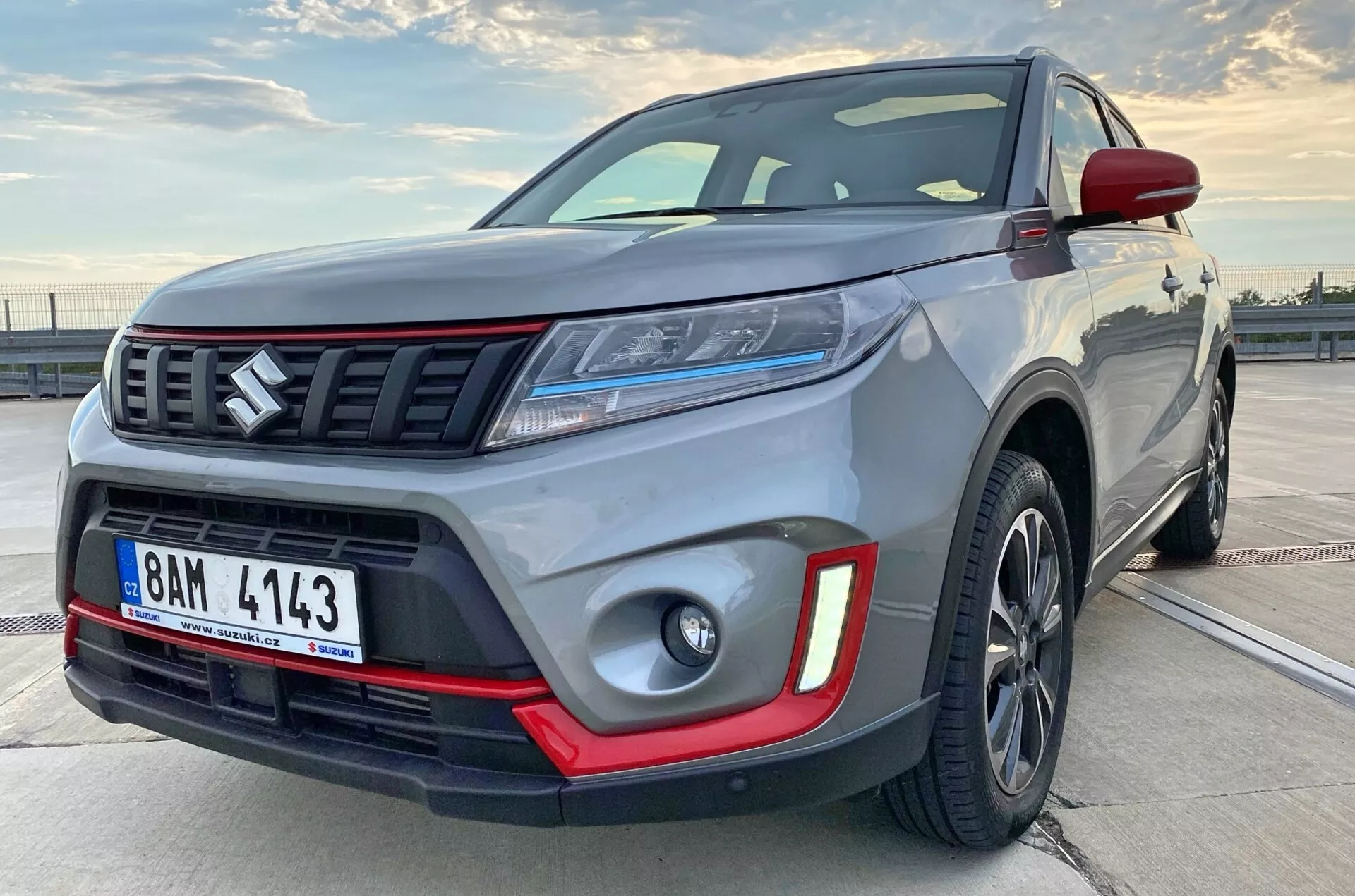 Närbild av en modern kompakt SUV från den främre sidovynkel, som betonar fordonets robusthet och storlek. Bilden visar detaljer som framstötfångaren, grillen med horisontella stänger och emblem, eleganta LED-strålkastare, dimljus med röda insatser, legeringshjul och muskulösa skärmar. Mörka aluminiumsfälgar och kontrasterande färgindikering pekar mot en premium- eller sportversion.
