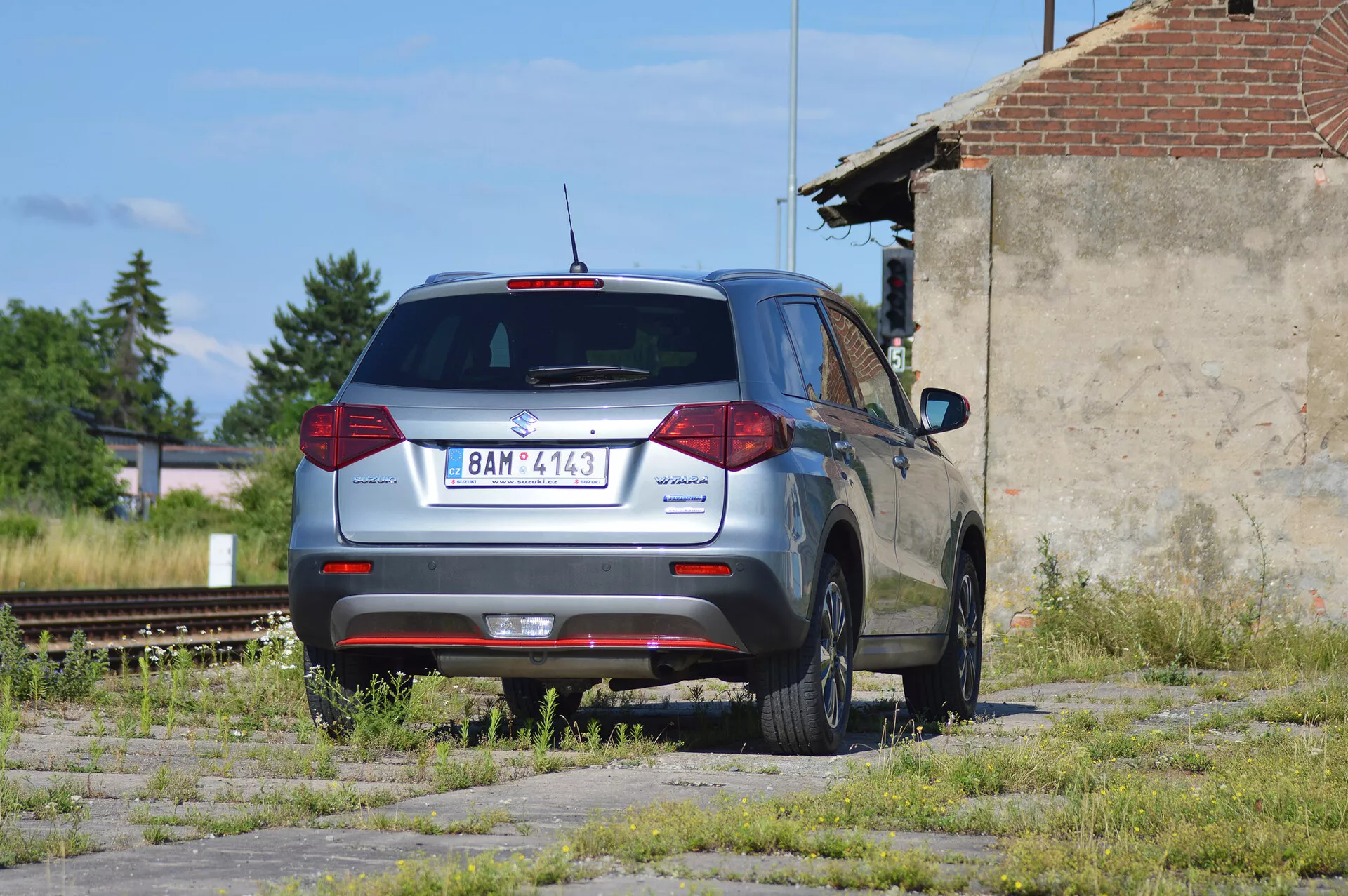 En metallicgrå Škoda Yeti kompakt SUV parkerad på en förfallen tomt med fläckar av gräs och betong, fotograferad från en bakre trekvarts vy. Bilden visar fordonets distinkta bakre design med konventionella bakljus, bakre vindrutetorkare, högt placerat bromsljus och en delvis synlig bakspoiler. Škoda-loggan är centralt placerad på bakluckan med modellnamnet 'Yeti' till vänster och en '4x4' badge till höger, vilket anger fyrhjulsdrift. Bilen har femekrade aluminiumfälgar, ser ut att vara i gott skick och är placerad i ett industriområde med nedlagda järnvägsspår och en förfallen struktur i bakgrunden, under naturligt solljus.