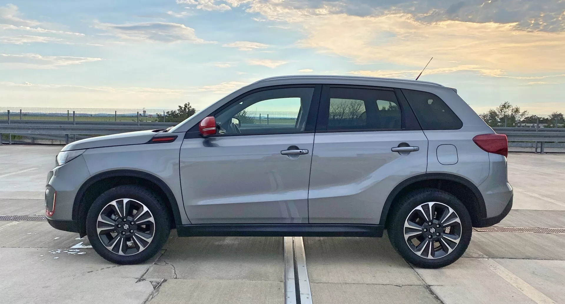 Sidoprofilbild av en modern SUV, Suzuki Vitara 2018, tagen under dagsljus på en rymlig parkering. Fokus ligger på fordonets mittsektion, vilket visar längden och sidadesign-elementen som dörrformer, sidotrösklar och hjulbågar. Det naturliga ljuset framhäver dess fem dörrar, flerspoksfälgar i lättmetall, takräcken, kontrasterande handtag och trimdelar, antenn och möjligen tonade fönster. SUV:en ser ut att vara i perfekt skick med en reflekterande glans på exteriören.