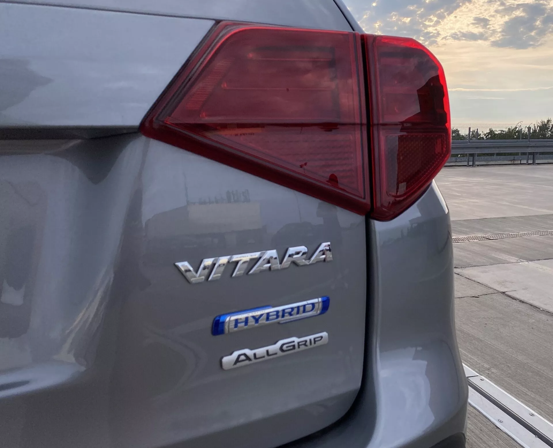 Närbild på den bakre vänstra delen av en Suzuki Vitara 2018 som visar dess intrikata LED-bakljusdesign och tre märken som indikerar modellnamnet 'VITARA', hybridteknik 'HYBRID' och fyrhjulsdriftsystemet 'ALLGRIP', fångat i gryning eller skymning på en öppen parkeringsplats.