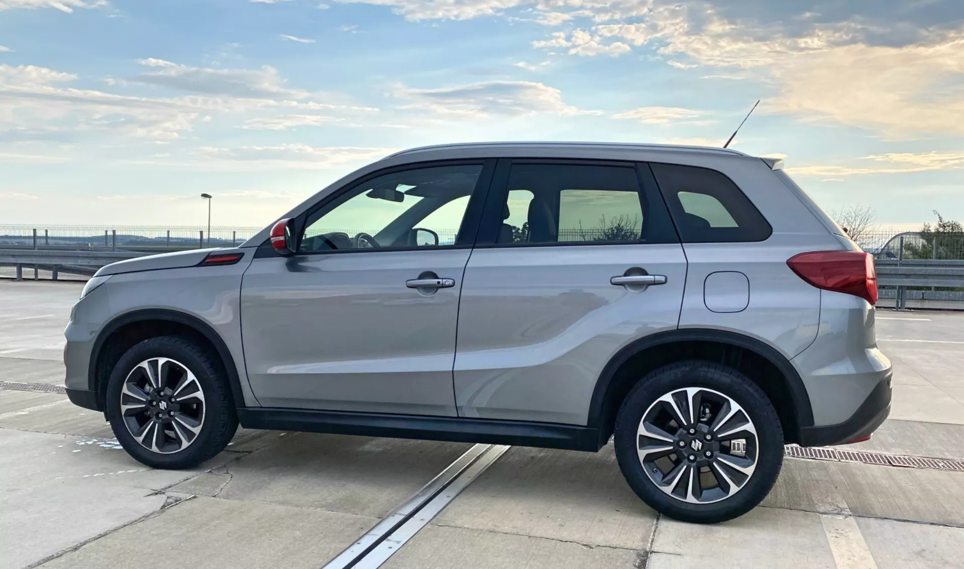 Sidoprofilvy av en Suzuki Vitara 2018 parkerad på det övre däck av en parkeringsstruktur under en klar himmel. SUV:en har femekrade lättmetallfälgar, silverlackering, integrerade blinkers på sidospeglarna, takräcken, dörrhandtag i karossens färg, en subtil sidostigbräda och tonade fönster med kromram. Fordonets design betonar sportig elegans och funktionalitet, med en kantig kaross för att maximera interiörutrymmet. Utomhusinställningen och kompositionen framhäver bilens stil och konstruktion, lämplig för fordonsrelaterade kategorier.