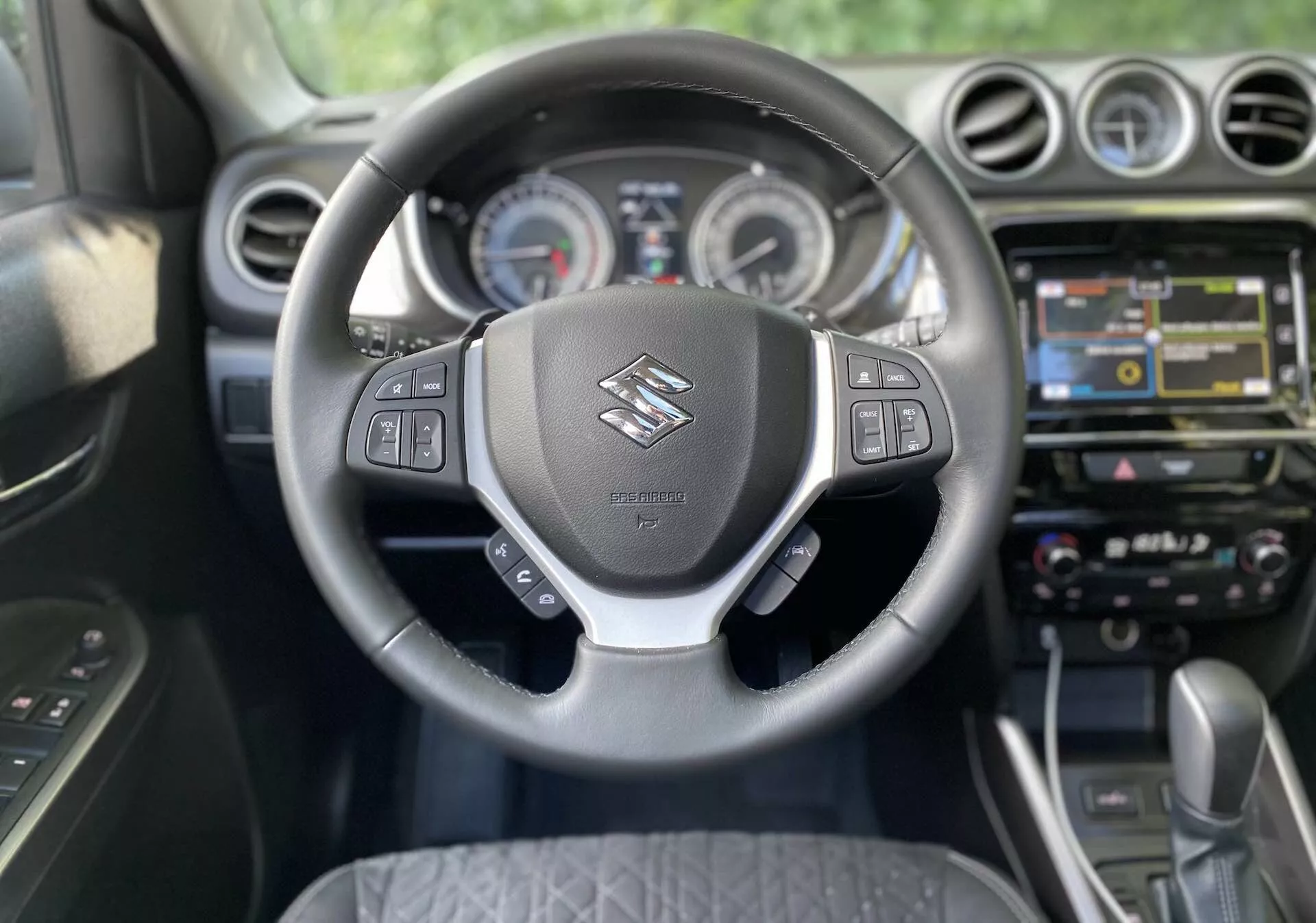 Interiörvy av en Suzuki Vitara 2018 från förarsätets perspektiv. Fokuset ligger på ratten, som har Suzuki-loggan och multifunktionskontroller för ljud, röstkommandon, farthållare och fordonets informationsdisplay. Bakom ratten syns analoga mätare och en central display på instrumentbrädan. Mittkonsolen visar ett infotainmentsystem med pekskärm med navigering, multimedia och anslutningsmöjligheter. Nedanför finns taktila knappar och vred för tvåzons klimatanläggning. Bilen är utrustad med en automatisk växellåda, vilket indikerar en modern och eventuellt premium kabindesign.