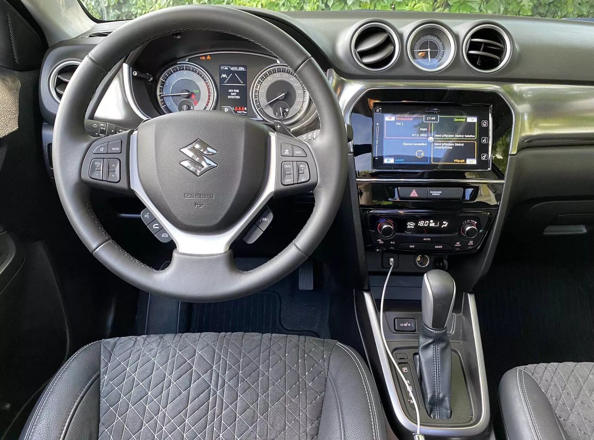 Interiörvy av en 2018 Suzuki Vitara från förarsätet, som visar det centrala ratthjulet med varumärkesloggan, kontrollknappar för volym, röstkommando och telefonanslutning. Bakom ratten finns en digital-analog instrumentpanel med hastighetsmätare, varvräknare och en central digital display. Instrumentbrädan har en pekskärms-infotainment med GPS-navigering, radio och klimatanläggning. Den automatiska växelspaken och den elektroniska parkeringsbromsen är tydligt synliga, tillsammans med en quiltad sätesdesign och cirkulära luftventiler, som fångar modern bilteknik och ergonomisk design.