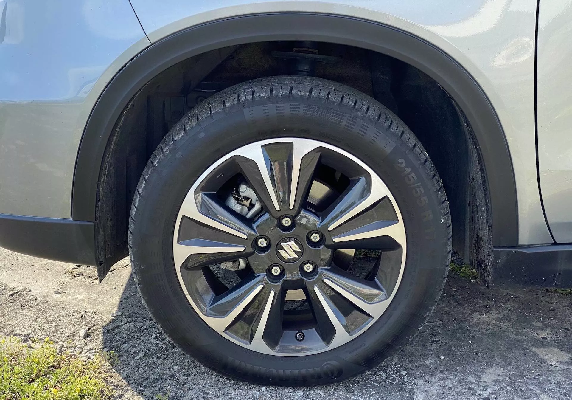 Närbild av hjulet på en Suzuki Vitara 2018, som visar en lättmetallfälg med flera metalliska och mörkare målade ekrar. Det märkesförsedda centrala locket döljer hjulmutter och nav, och däcket för vägbruk har detaljerade mönster och sidoväggsmärkningar. Bakgrunden inkluderar en del av karossens skärm och sidopanel i en neutral och glansig färg, sett från en något uppåtriktad vinkel på marknivå.