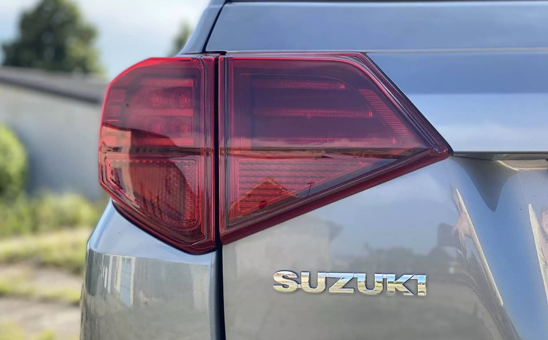 Närbild av den bakre vänstra sidan av en Suzuki Vitara 2018 med fokus på baklyktan. Perspektivbild tagen från låg till medelhög höjd och som visar tydligt utformad LED-baklykta med skarpa vinklar och flera kammare för funktioner som bromsljus, blinkers och eventuellt dimljus. Den främst röda baklyktan med en genomskinlig sektion, troligen för backljuset, framhäver modern fordonsbelysningsteknik. Bakluckan har en centralt placerad kromad 'SUZUKI' logotyp under bakrutan som betonar ren och jämn märkning. Den metalliska lacken reflekterar lite ljus och indikerar en väl underhållen eller nyare yta på fordonet.