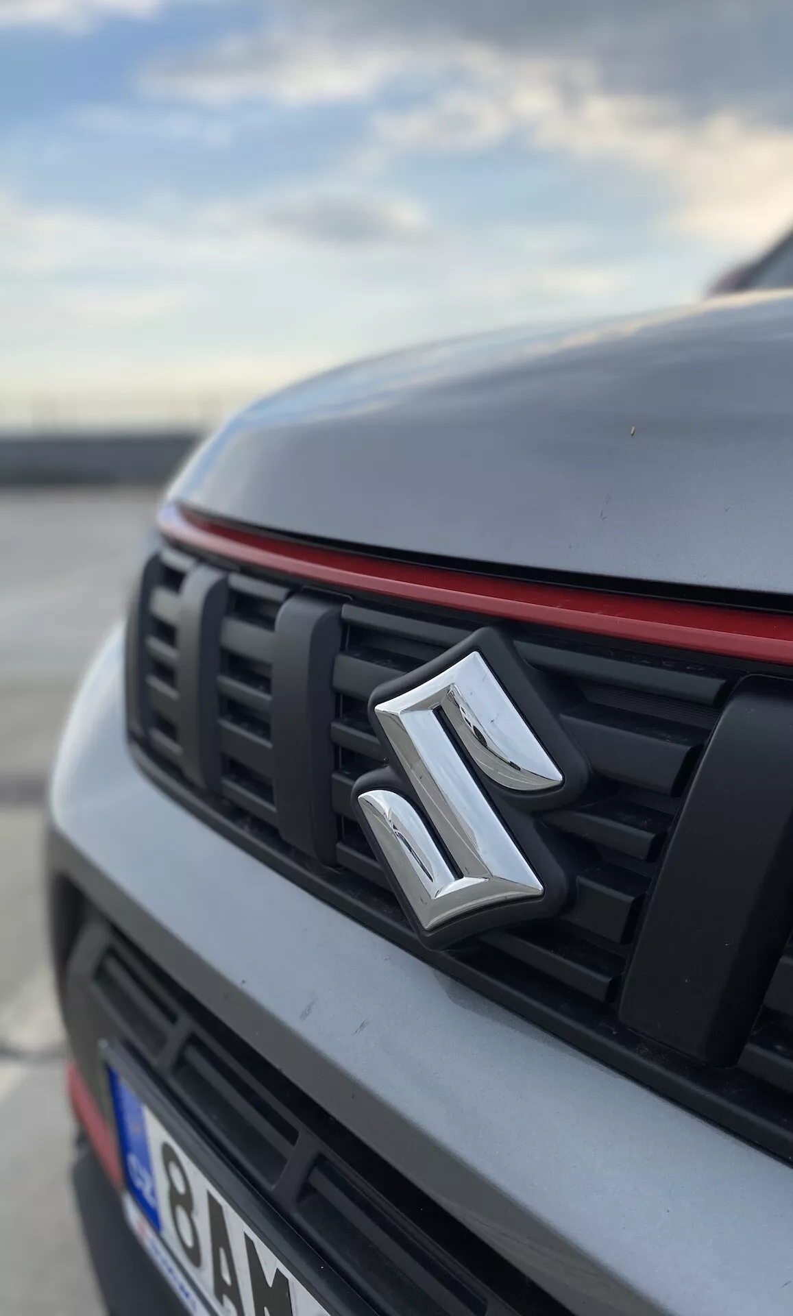 Närbild, låg vinkelvy av fronten på en Suzuki Vitara 2018. Bilden framhäver tillverkarens emblem centrerat på gallret som har horisontella slatsar och en röd kant längs överkanten vilket antyder en sportigare design. Huven och delar av bilen är suddiga vilket framhäver en vit eller ljus finish med ett urskiljbart veck. Bakgrunden är också suddig och antyder en utomhusmiljö, möjligen på en parkeringsplats, under tidig morgon eller sen eftermiddag.