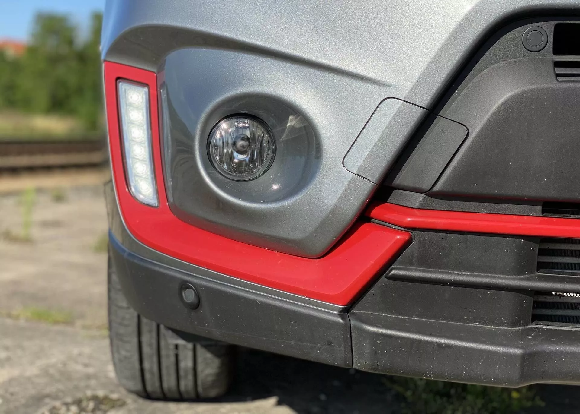 Närbild av bakre hörnet på en Suzuki Vitara 2018, med en klarlinsad dimlampa i bakre stötfångaren, en modern kantig design på baklyktorna, tvåfärgad bakre stötfångare med en sportig röd accent, potentiella parkeringssensorer, lågprofildäck med skyddande plast hjulbåge, blank färgfinish och en oskarp halvindustriell bakgrund med järnvägsspår.