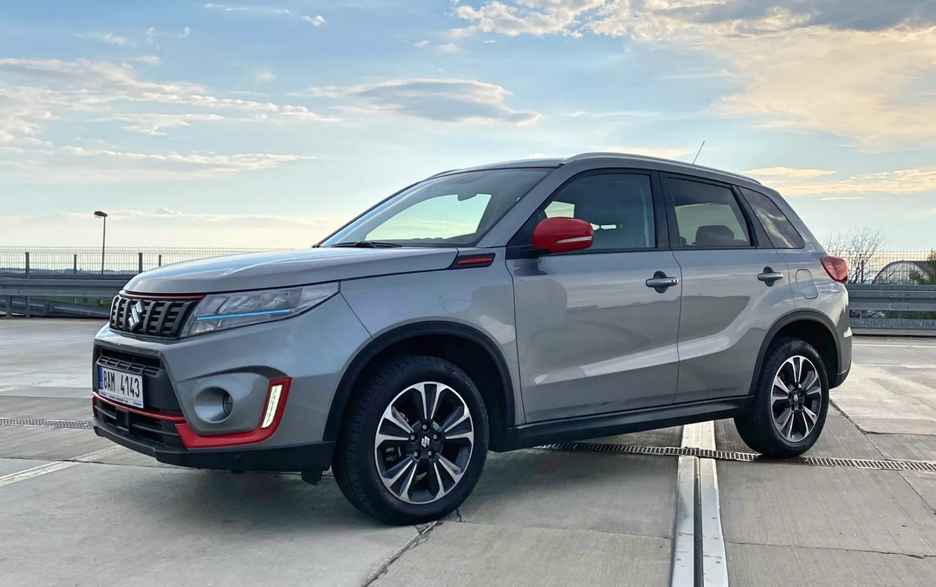 Bilden visar en Suzuki Vitara 2018, en modern kompaktsuv parkad utomhus i dagsljus. Den är fotograferad i en trekvarts framifrånvy som visar både fronten och sidoprofilen. Vinkeln är något låg, i nivå med dörrhandtagen, vilket antyder att fotografen hukade eller böjde sig. Synliga detaljer inkluderar frontgrillen, strålkastarna, dimljushus, emblem, sidospeglar, lättmetallfälgar med integrerade blinkers, dörrhandtag, fönsterlister, taklinje och takräcken. Projektorlampor och elegant antenn indikerar avancerad teknik. De moderna designelementen hos SUV:en indikerar funktioner som LED-lampor och möjligtvis ett nyckelfritt ingångssystem.