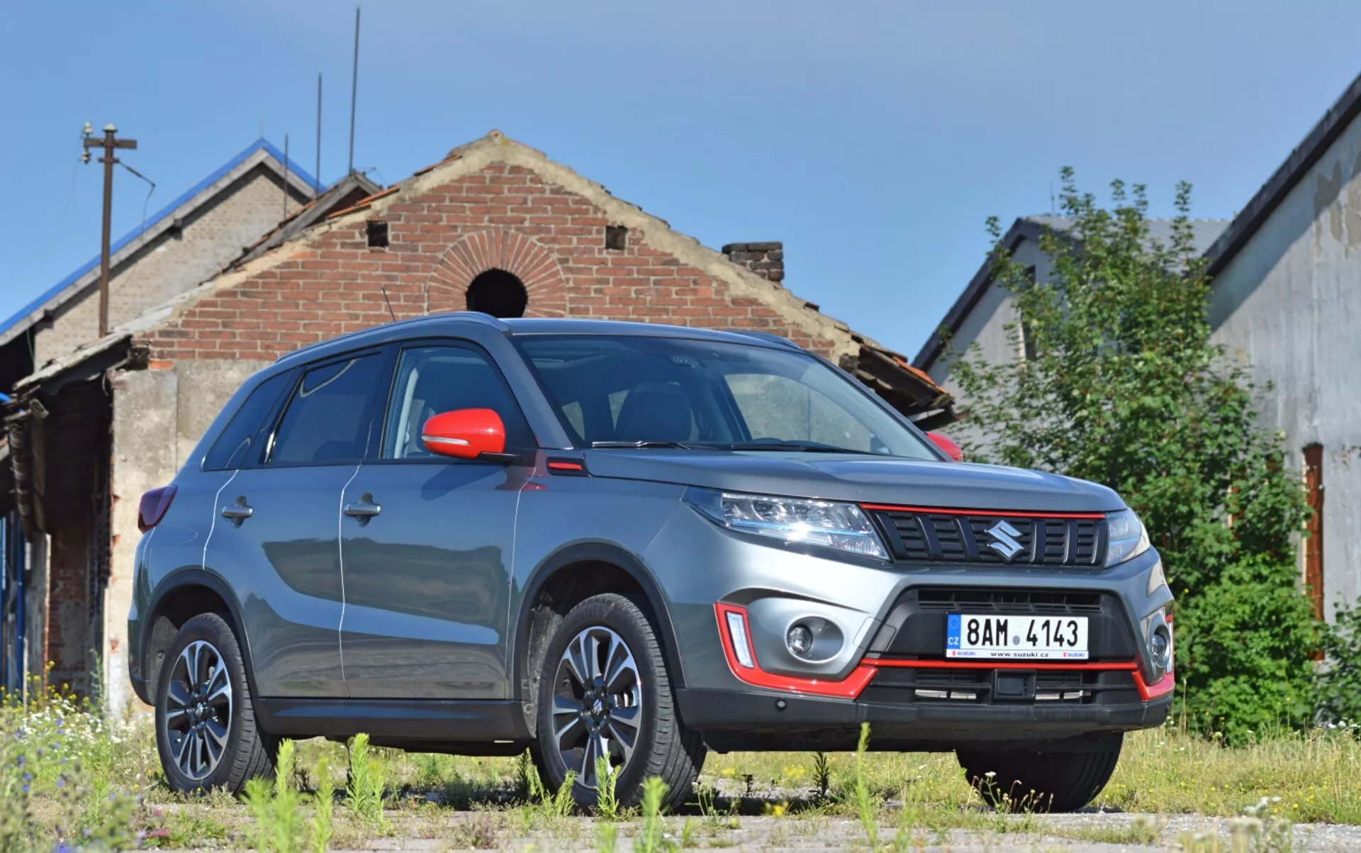 Bilden visar en 2018 Suzuki Vitara SUV från en låg trekvarts framifrån vy, vilket framhäver fordonets framträdande högra framdel med märkbar markfrigång. Synliga funktioner inkluderar en robust främre stötfångare med dimljus och silverfärgad släpskydd, varumärkets emblem på den horisontella grillen, definierade motorhuvslinjer, moderna strålkastare med möjlig LED- eller HID-teknik, flerspoks legeringshjul, röda sidobackspeglar med integrerade blinkers, muskulösa skärmar, reflekterande vindruta och tonade fönster. Vitaran är parkerad på en ojämn gräsyta, framför en gammal byggnad som ger en industriell eller lantlig bakgrund, vilket antyder lämplighet för utomhusmiljöer.