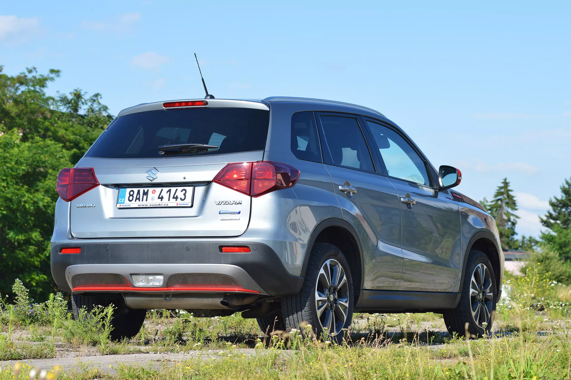 2018 Suzuki Vitara fångad från en trekvarts bakre höger vinkel, som betonar bakljusdesignen, bakrutan och passagerarsidans profil. Bilden visar den kompakt SUV:ns moderna bekvämligheter som en takantenn för teknologiska funktioner, bakre parkeringssensorer och stiliga lättmetallfälgar. Fordonet är placerat på en ojämn yta i lantlig miljö, vilket indikerar dess terrängmöjligheter. Idealisk för fordons-, SUV- och terrängbilskategorier.