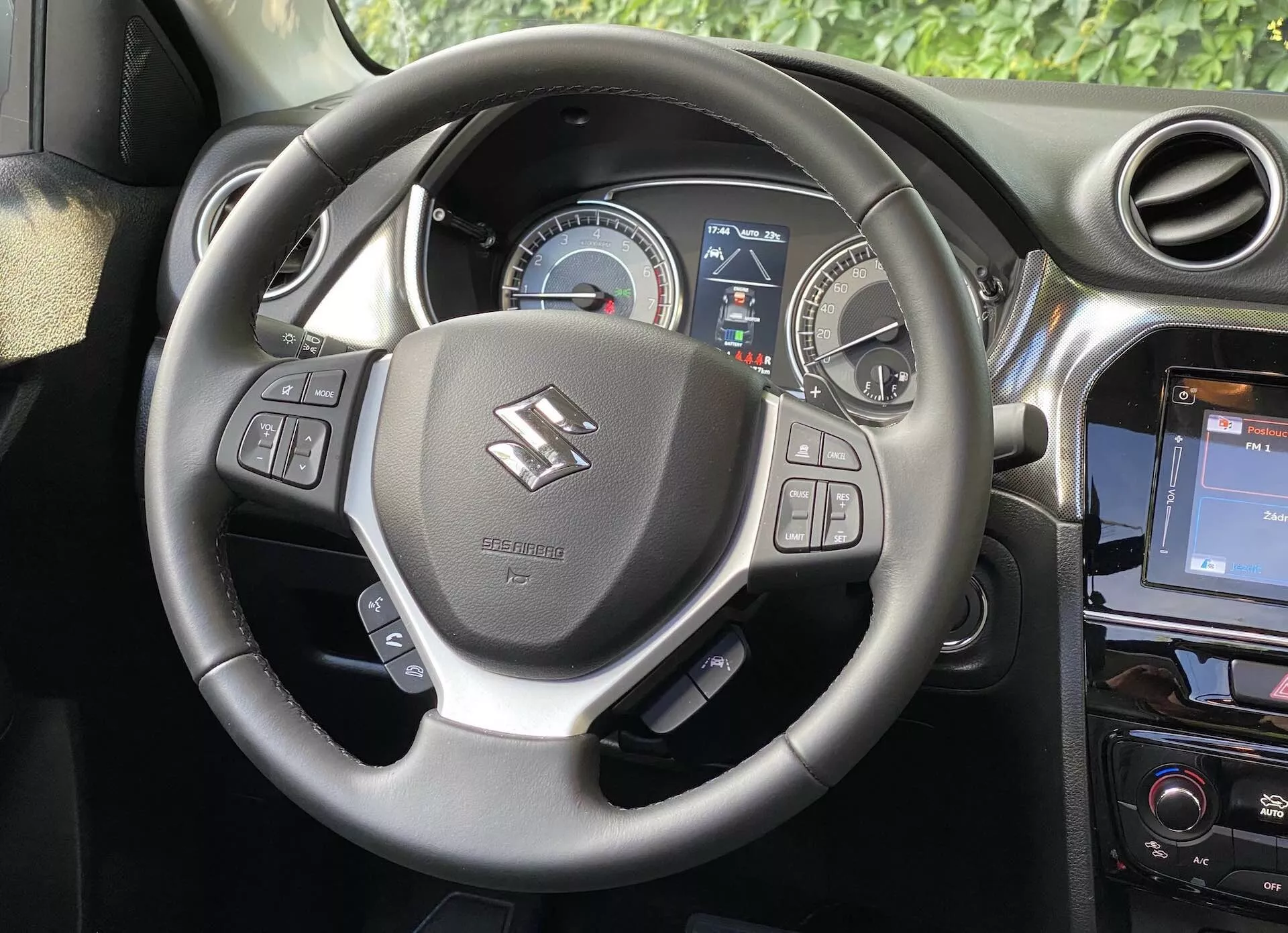 Interiörbild av en Suzuki Vitara 2018, som visar förarens perspektiv med fokus på ratt och instrumentbräda. Ratten, som är centrerad i bilden, har tumstöd och kontroller för ljud, röstkommandon och farthållare. Bakom den finns en instrumentpanel med en analog hastighetsmätare till höger och en delvis digital display som ger förarinformation till vänster. En del av mittkonsolen till höger visar ett pekskärmsinformationssystem, vilket förstärker bilens moderna estetik.