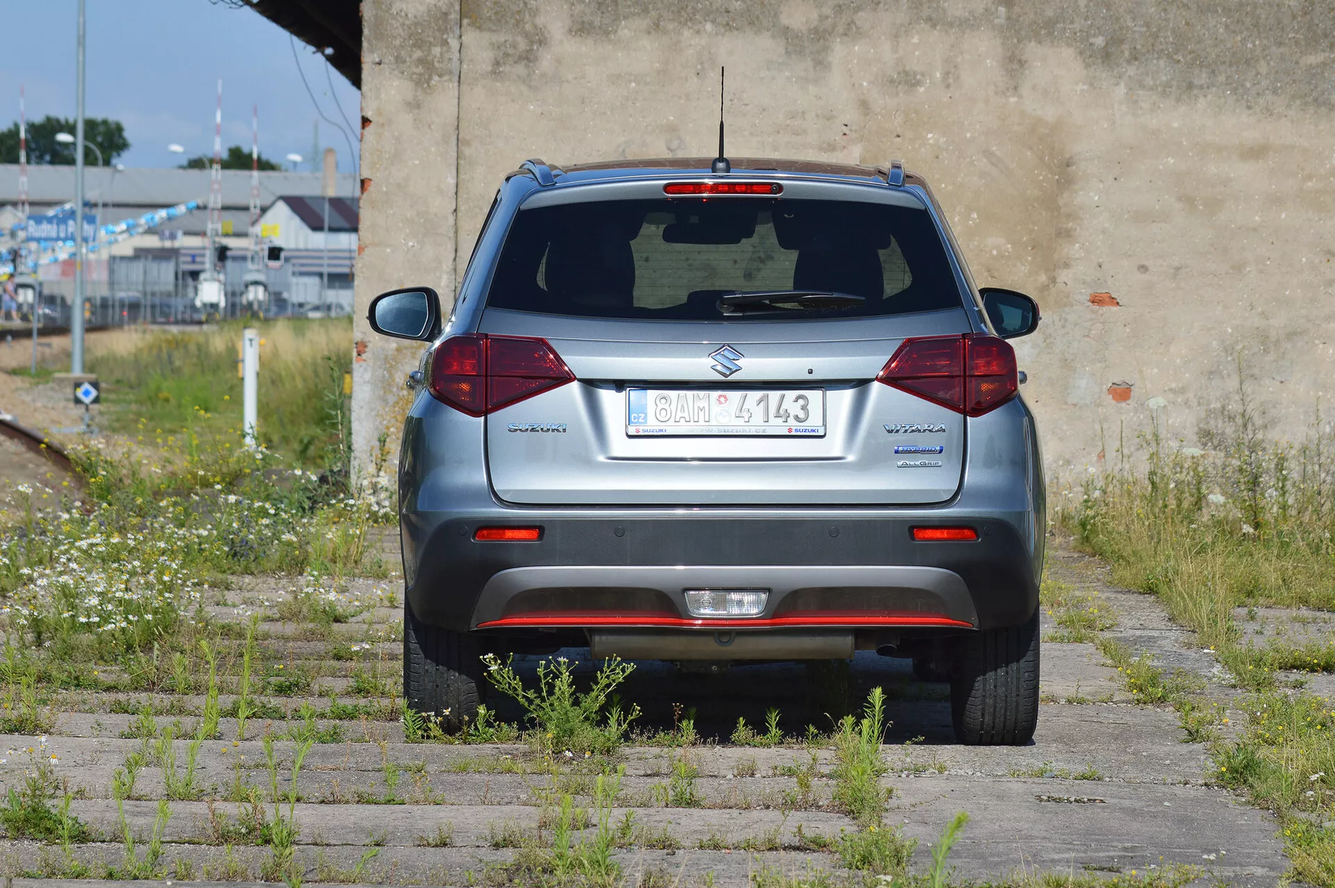 Bakvinkelsbild av en silverfärgad kompakt SUV Suzuki Vitara 2018 parkerad på gräs och betongplattor. Hela den bakre sektionen är synlig, med en centrerad registreringsskylt, bakruteviskare, tredje bromsljus, standard halogenbakljus, reflexer på stötfångaren, parkeringssensorer och ett avgasrör på vänster sida. Fordonet befinner sig i ett halvövergivet område med övervuxen vegetation och en väderbiten vägg.