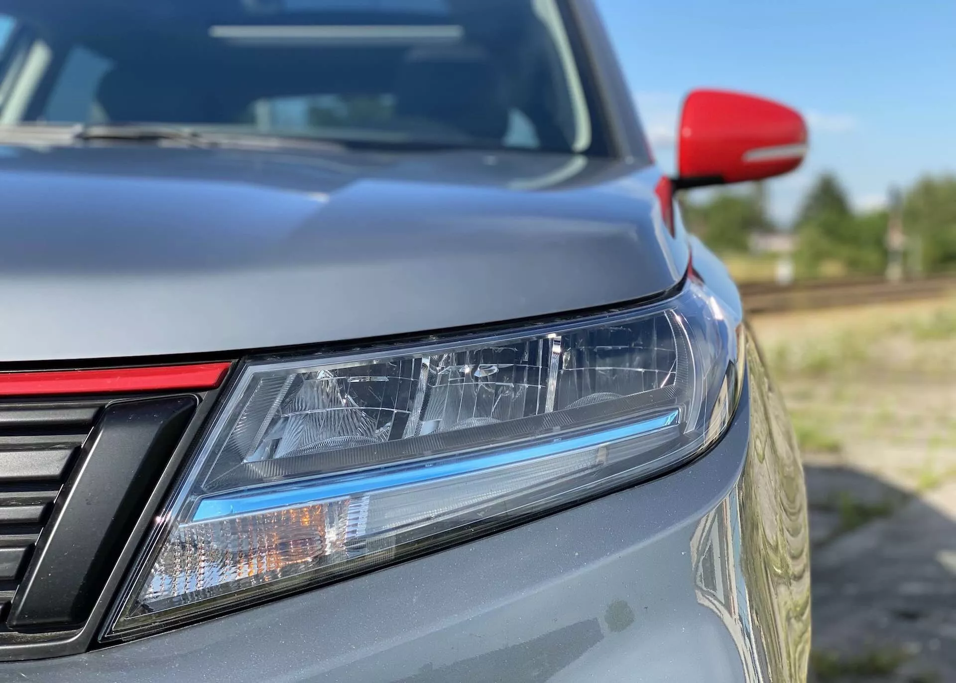 Närbild av vänstra främre hörnet på en 2018 Suzuki Vitara, med fokus på detaljerade funktioner som LED-strålkastare med dagsljusljus, en delvis synlig grill med vertikala ribbor, en mjukt sluttande motorhuv, metallisk lack, en kontrasterande röd sidospegel troligen med integrerade blinkers och en oskarp bakgrund som framhäver den slanka designen och aerodynamiska effektiviteten.