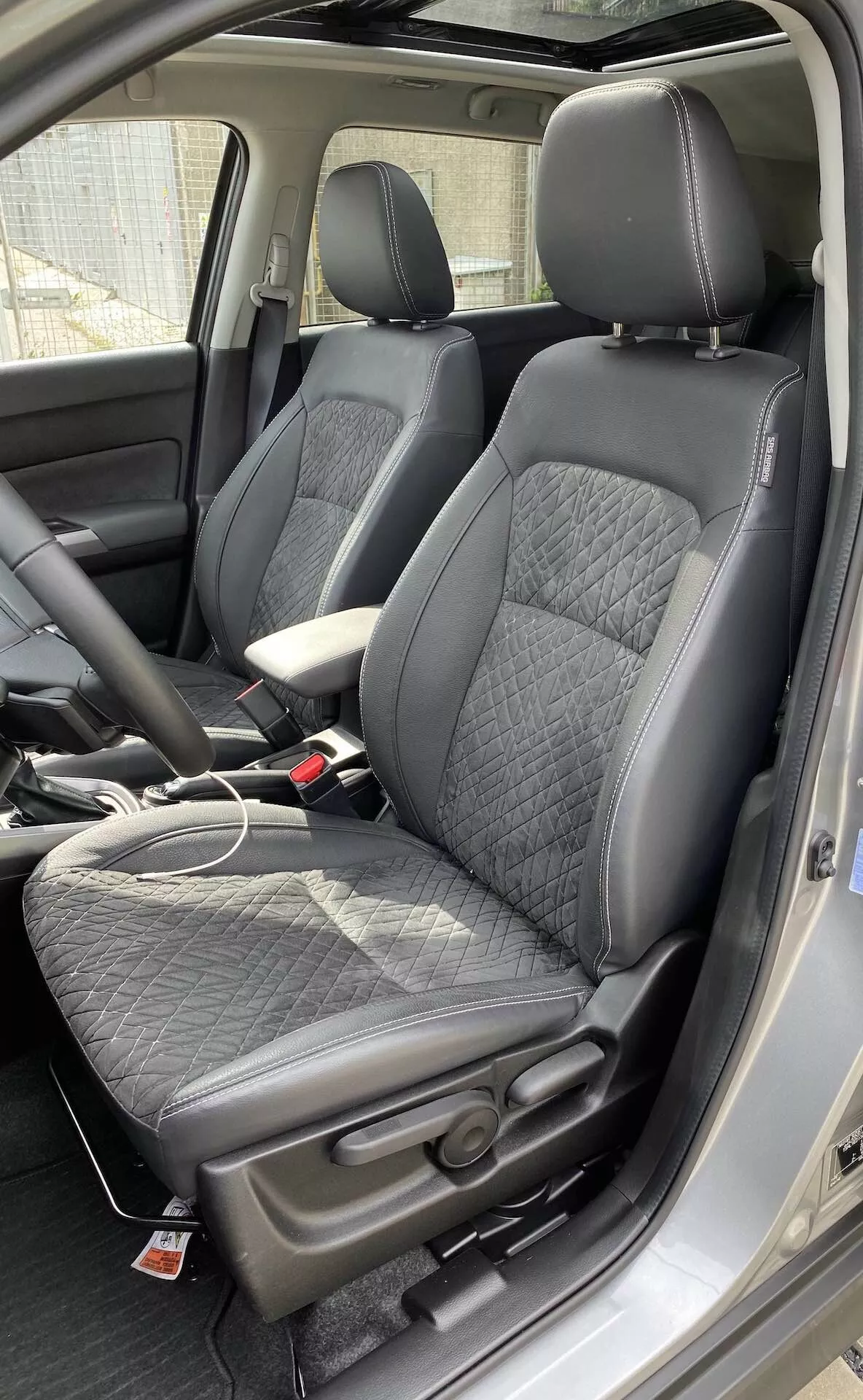 Interiören i en Suzuki Vitara 2018, som framhäver de moderna hinkstilsframsätena från en något ovanför sitsens perspektiv. Sätena är klädda i svart läder med ett diamantmönstrat quiltat insats och släta lädersidor, vilket erbjuder en balans mellan komfort och sportighet. En röd etikett på sätena antyder ytterligare funktioner som sidokrockkuddar eller uppvärmda säten. Den centrala konsolen med ett läderbeklätt armstöd är synlig, liksom en laddkabel på passagerarsätet, vilket indikerar närvaron av laddningsportar. Säkerhetsbältesspännena matchar det övergripande svarta interiörfärgschemat. En del av instrumentbrädan och B-stolpen är också synliga, vilket antyder en rymlig kabin.