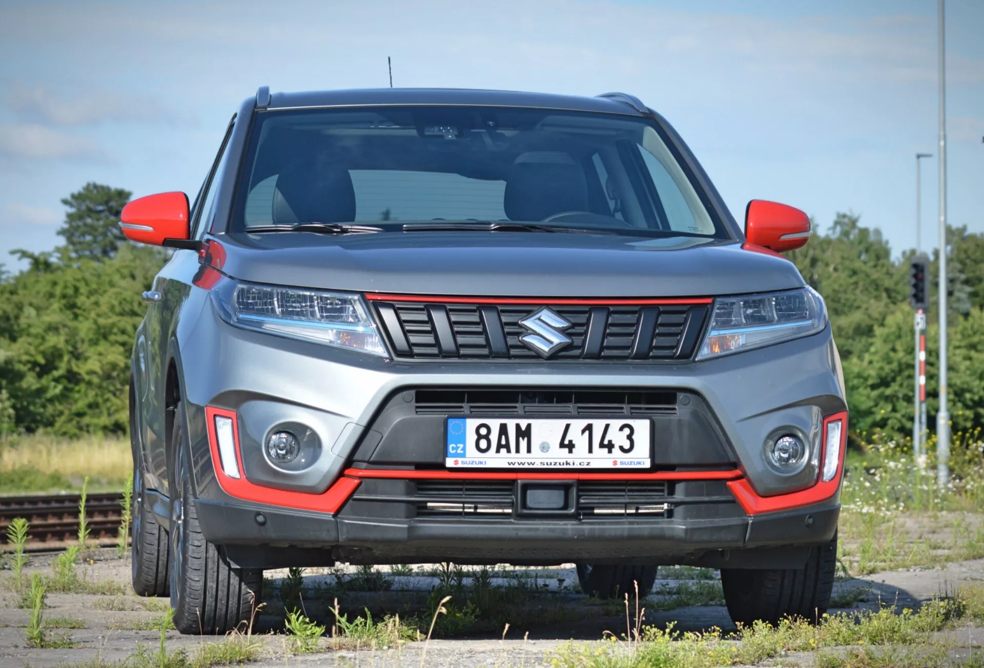 Bilden visar en modern, 2018 Suzuki Vitara SUV från en något främre vinkel, vilket betonar frontpartiet och en del av passagerarsidan mot en bakgrund av ett sparsmakat landskap med järnvägsspår. Bilen, parkerad på vad som verkar vara en grusyta, har en framträdande grill med en vinkelböjd design, eleganta strålkastare sannolikt med LED- eller HID-teknik, och integrerade dagsljuslampor eller dimljus i främre stötfångaren. Stötfångaren innehåller en kontrasterande färginsats, vilket tyder på en terräng- eller sportig trimnivå och den synliga markfrigången förstärker dess SUV-karaktär. Upphöjda linjer på huven bidrar till en aggressiv design, medan sidospeglarna och framskärmarna visar en modern design. De delvis synliga lättmetallfälgarna kompletterar det robusta utseendet. Fotot, taget på nära håll, möjliggör en detaljerad granskning av dessa funktioner.