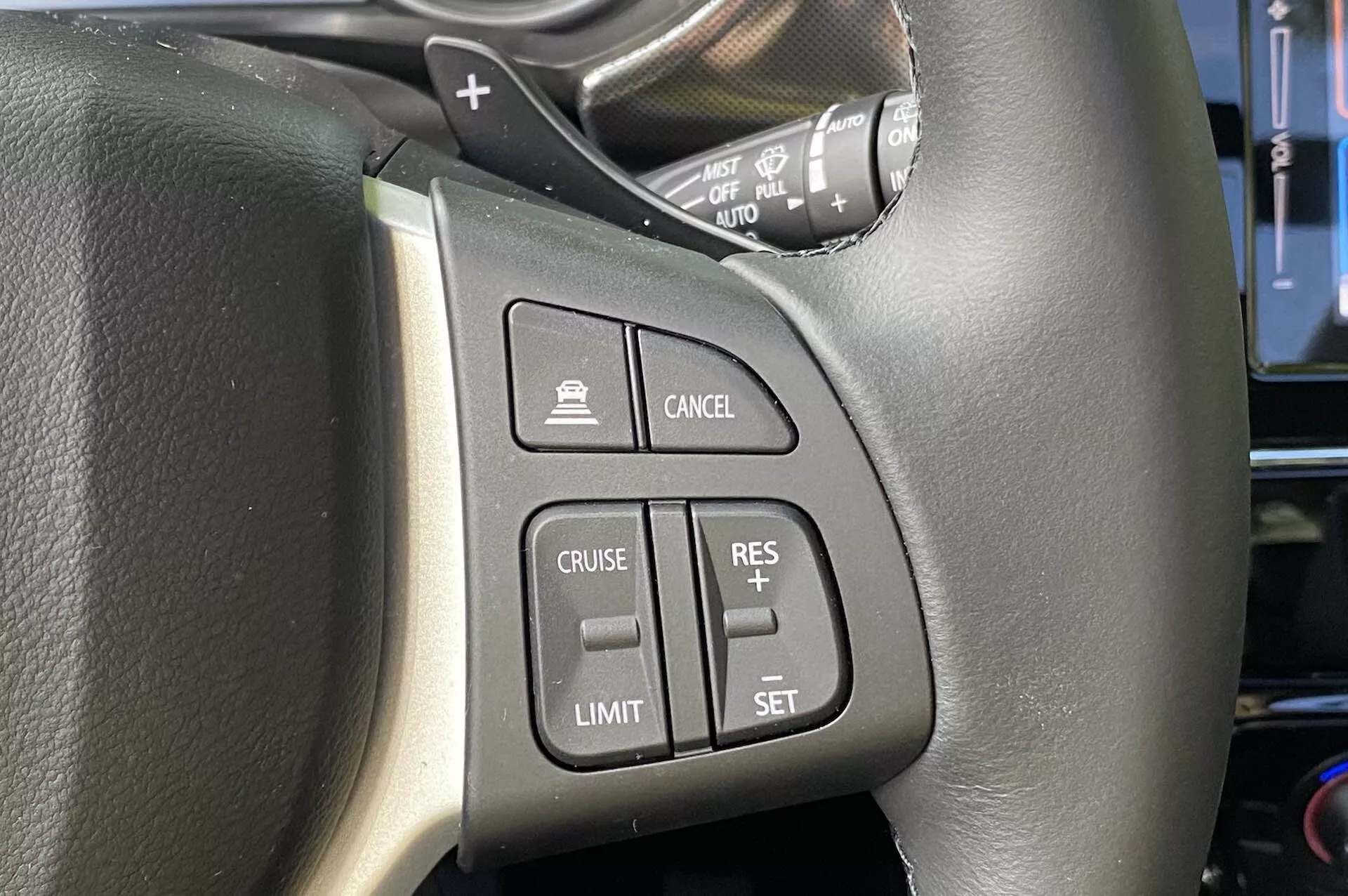 Närbild av rattmonterade reglage i Suzuki Vitara 2018, med fokus på vänster sida. Den svarta läderratt med synliga sömmar har lättillgängliga knappar inklusive aktivering av farthållare, aktivering av hastighetsbegränsare, farthållarens inställningsjustering och avbrytknapp. Bilden visar också en spak som troligen är för blinkers och strålkastare, sedd ur förarens perspektiv.