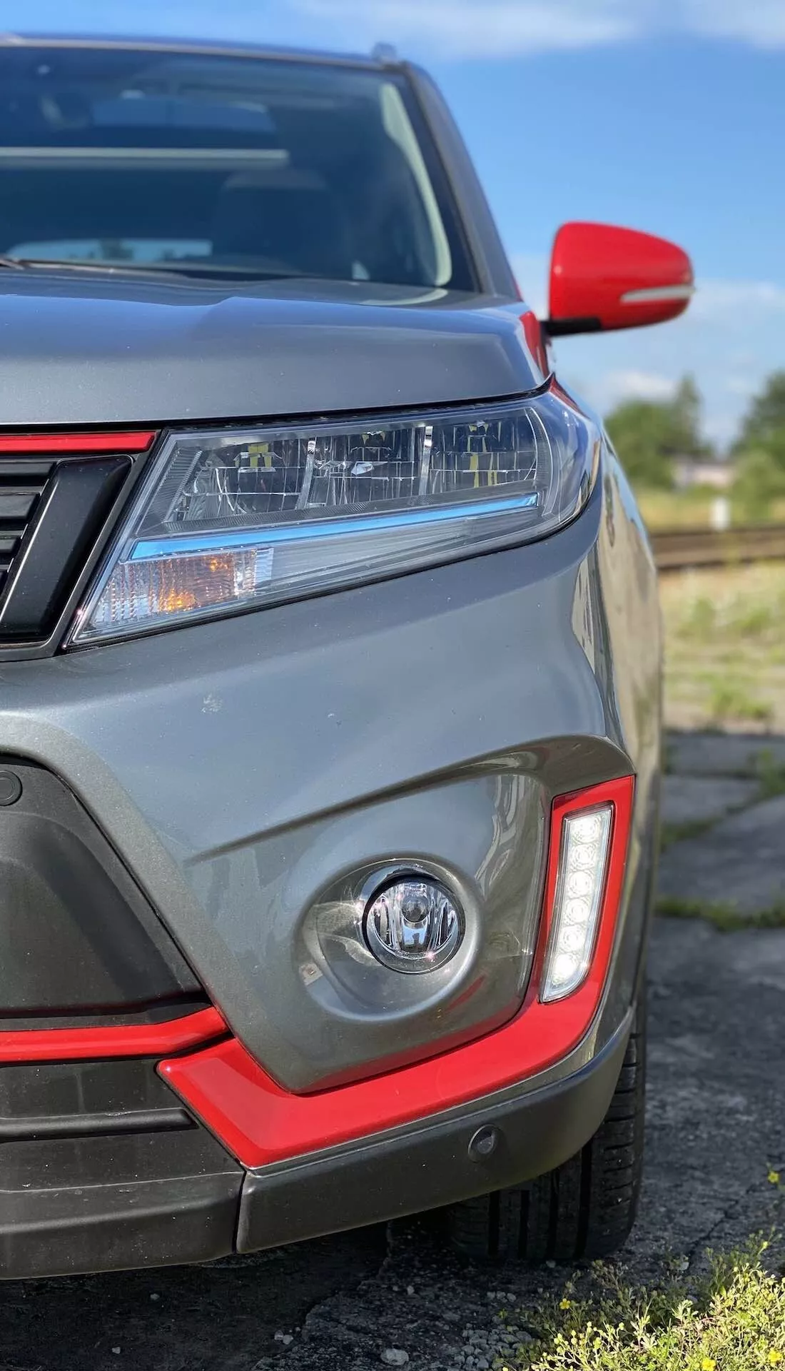 Närbild på den främre vänstra kvadranten av Suzuki Vitara 2018 som visar LED-strålkastare, dagsljuslampor, dimljus med röd accent, parkeringssensorer, delvis frontgrill, rödlockad sidospegel och metallisk grå färg.