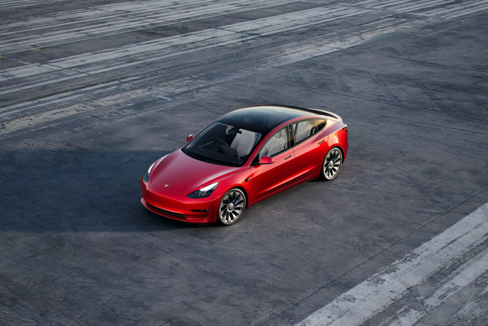Högvinkelsskott av en röd Tesla Model 3 (2019, 1:a generationen) fyrdörrars sedan parkerad på en texturerad asfaltyta med synliga däckmärken, vilket tyder på tidigare fordonsaktivitet. Bilen är något placerad till höger om bildens mitt och ses från ett trekvartsperspektiv framifrån, som visar förarsidan fram och en delvis bakre vy. Den aerodynamiska designen, med en distinkt konturlinje, panoramaglastak, fler ekrade lättmetallfälgar, LED-strålkastare och en minimalistisk frontgrill, lyfter fram fordonets avancerade elektriska teknik och design.