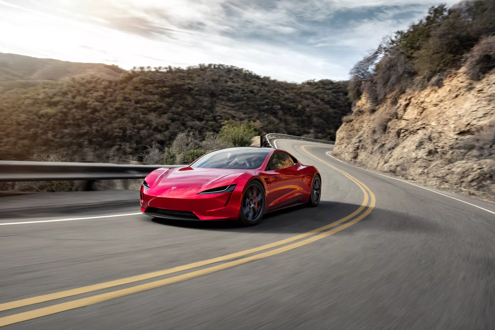 Bild av en ljusröd Tesla Roadster 2020 (2:a generationen) sportbil i rörelse på en slingrande väg. Trekvarts frontvy visar eleganta strålkastare, aerodynamisk grill, stora främre luftintag och formade linjer på huven. Bilens imponerande utseende förstärks av låg vinkel. Den suddiga vägen och bakgrunden antyder hög hastighet, med bilen placerad på vägens högra sida mot en bakgrund av klar himmel och växtlighet.