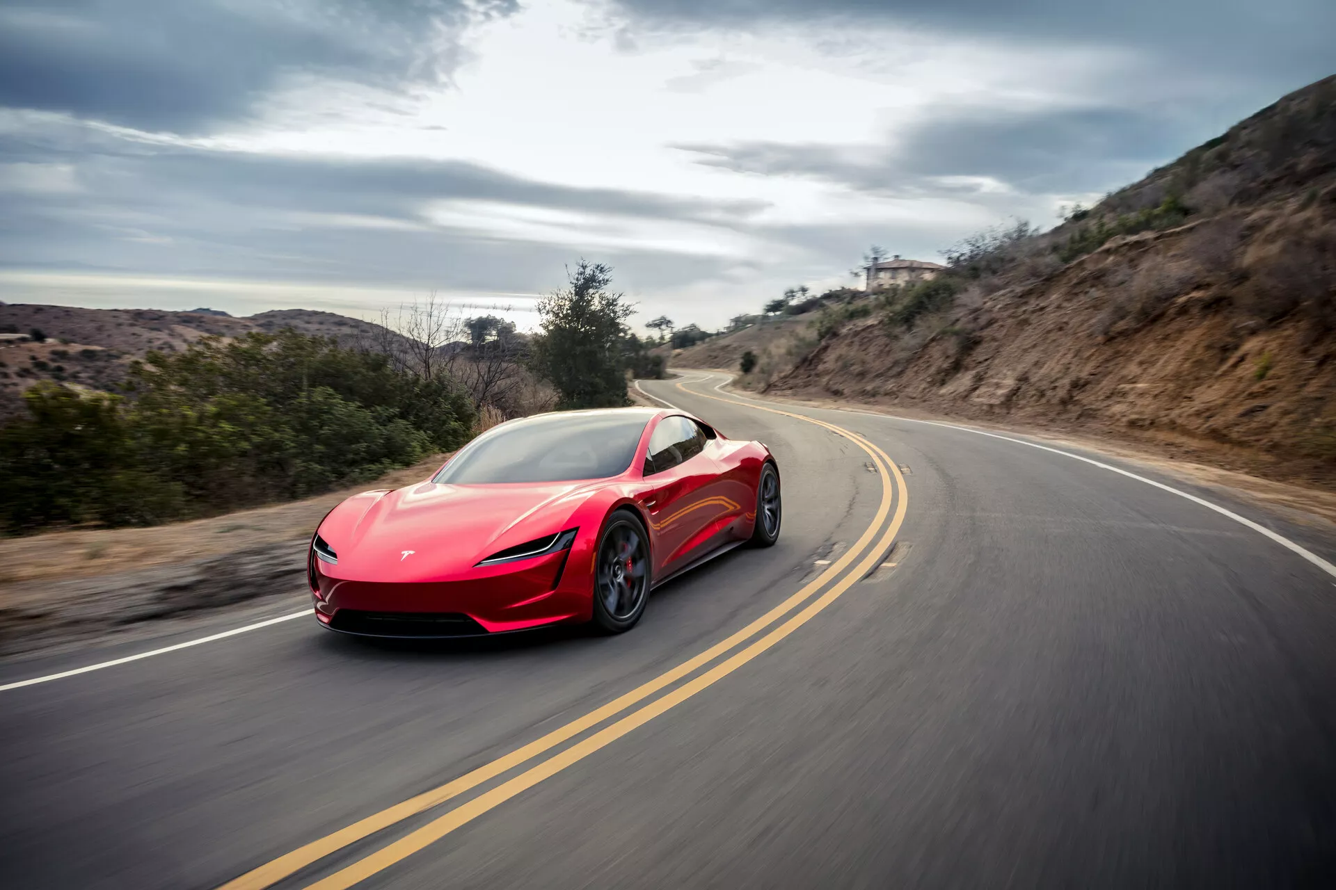 Dynamisk bild av en röd Tesla Roadster 2020 (andra generationen) sportbil fångad i rörelse på en slingrande väg som visar dess aerodynamiska linjer, eleganta strålkastare, sidospeglar, stora hjul och luftintag, mot en diffust lysande bakgrund som indikera höghastighetskörning i en lantlig miljö.