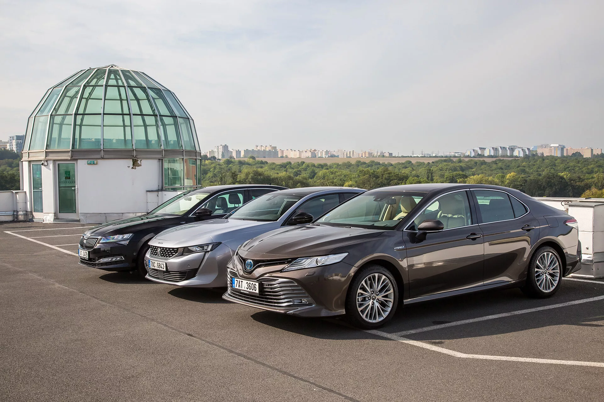 Toyota Camry Limousine från 2019 parkerad på takterass med klar himmel och glaskupol i bakgrunden, som visar en modern design med dynamisk frontgrill, vassa LED-strålkastare, skulpterad motorhuv och lyxiga aluminiumfälgar
