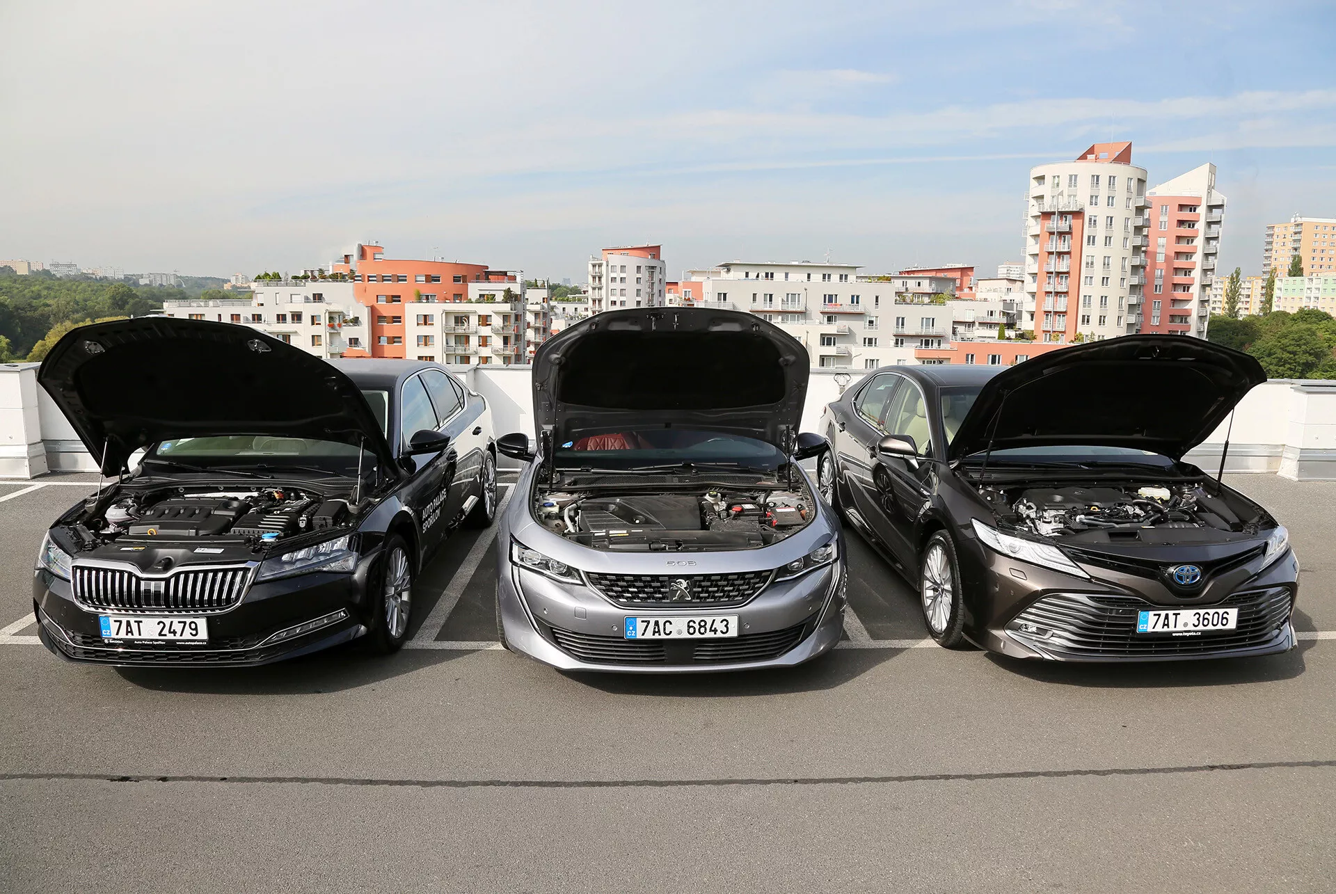 2019 Toyota Camry Limousine-modeller med öppna motorhuv som visar motorerna, parkerade sida vid sida på en förhöjd struktur, med stadssilhuetten i bakgrunden, vilket framhäver fordonens slanka, moderna design och avancerad motorteknologi.