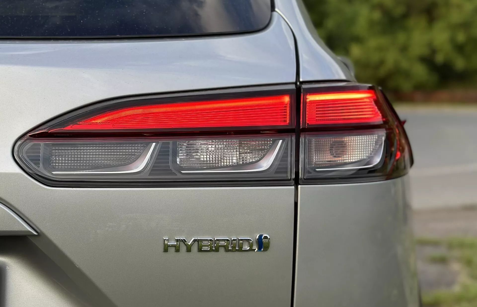 Närbild på vänstra bakre hörnet av Toyota Corolla-Cross 2022 som visar LED-baklyktedesign och HYBRID-emblem med fokus på fordonets miljövänliga teknologi och glänsande finish i en dämpad ljusgrå färg
