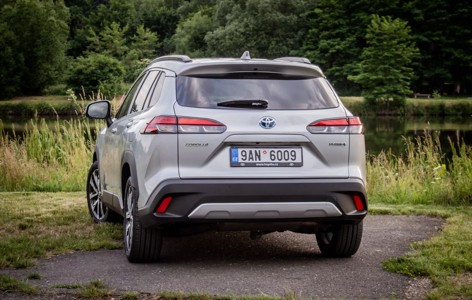 2022 silver Toyota Corolla Cross parkerad på ett trottoar med grönska i bakgrunden som visar dess LED-bakljus, bakre spoiler, flerspakig legeringshjul och hybridmärkning i naturligt dagsljus