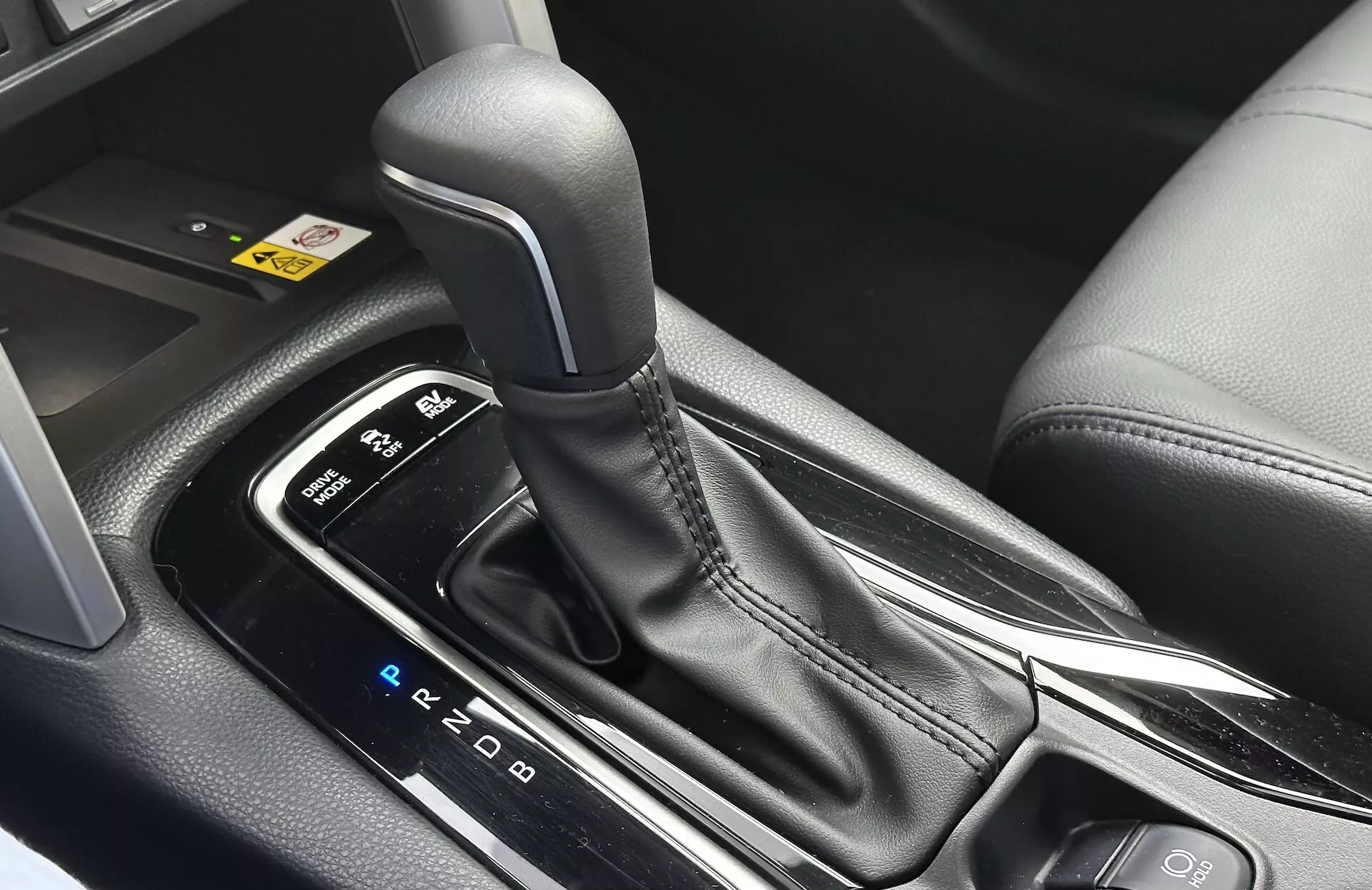Närbild på interiören i Toyota Corolla Cross 2022 som visar den automatiska växelspaksknoppens PRND-layout, omgiven av en elegant mittkonsol med svarta plast- och metallaccentdetaljer, framhävande ergonomisk design och moderna teknologiska funktioner