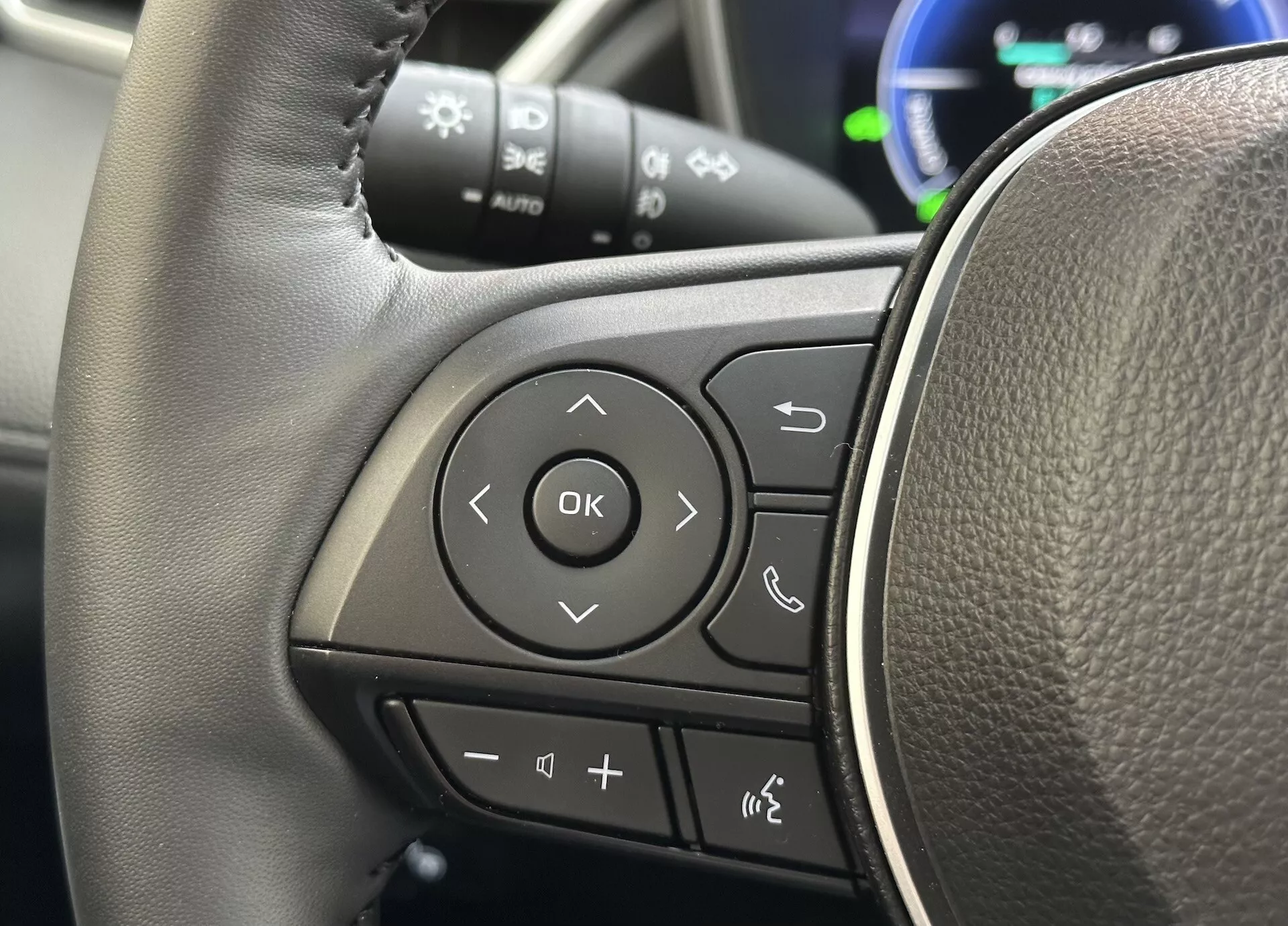 Detaljerad vy av rattet på Toyota Corolla Cross 2022 med fokus på multifunktionella kontroller på vänster ekrad med en läderliknande textur och tydligt markerade knappar för ljud, Bluetooth-telefonanslutning, röstkommandon och navigering genom förarens informationssystem